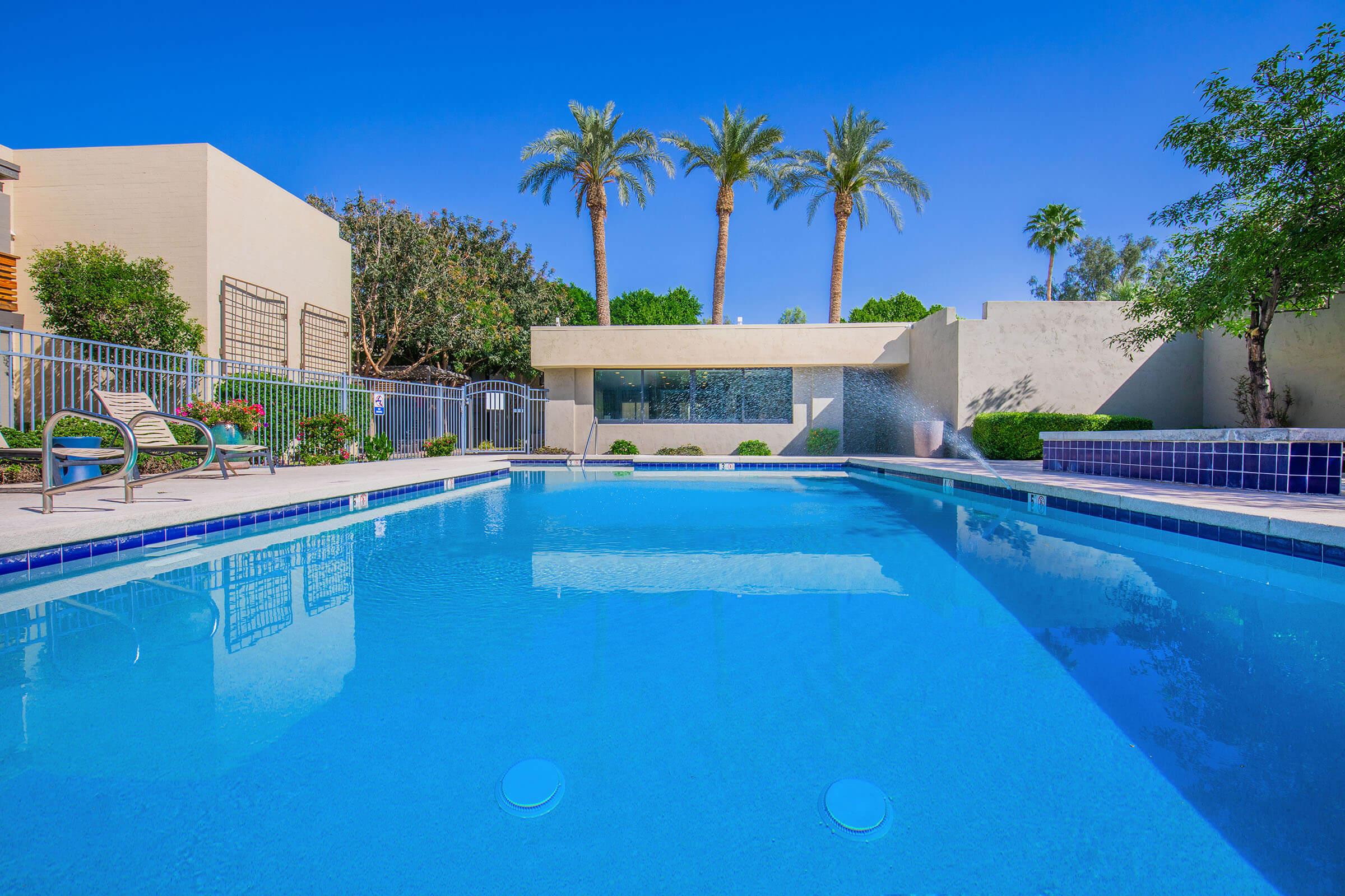 A serene outdoor swimming pool area surrounded by lush greenery and palm trees under a clear blue sky. The pool features a sleek design with a tiled edge and a small waterfall feature, inviting relaxation and leisure.