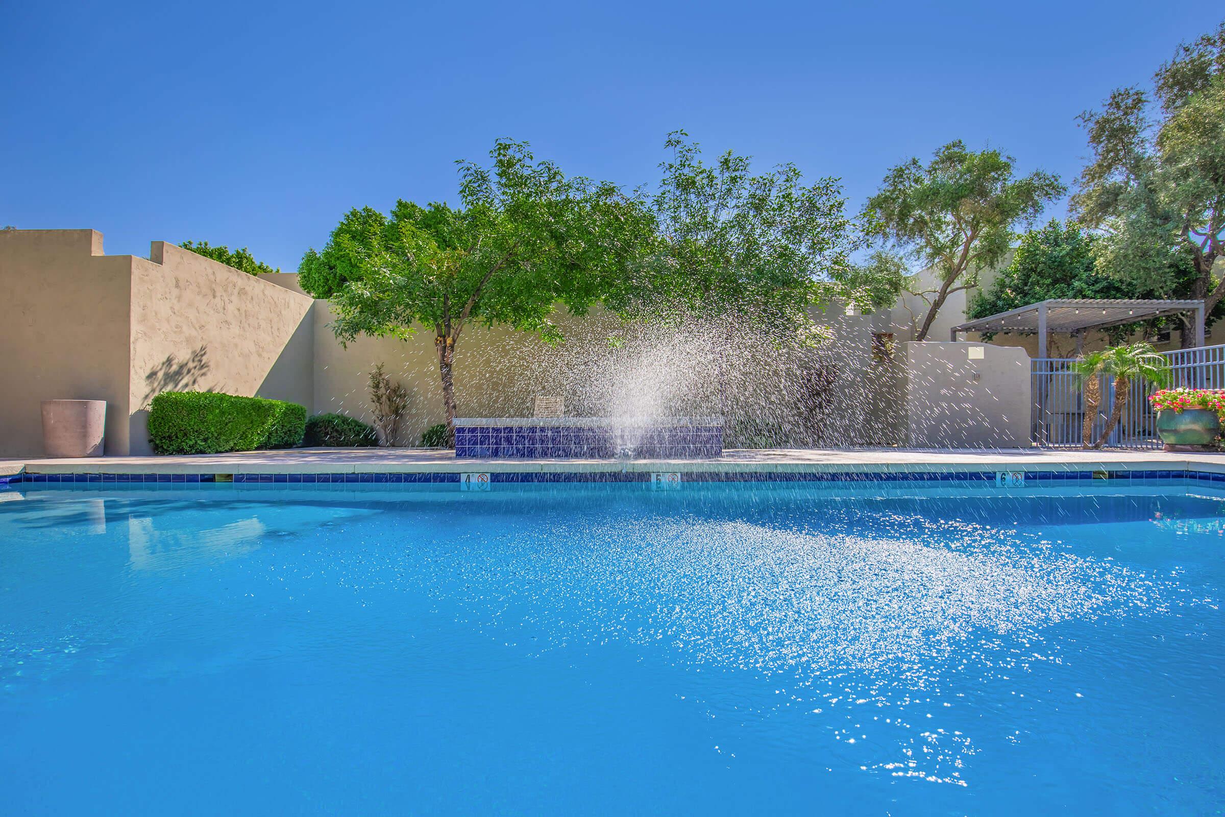 A clear swimming pool with water splashing from a fountain feature. Surrounding the pool are lush green trees and shrubs, with a sunny blue sky overhead. The scene is relaxing and inviting, suggesting a tranquil outdoor space ideal for leisure and enjoyment.