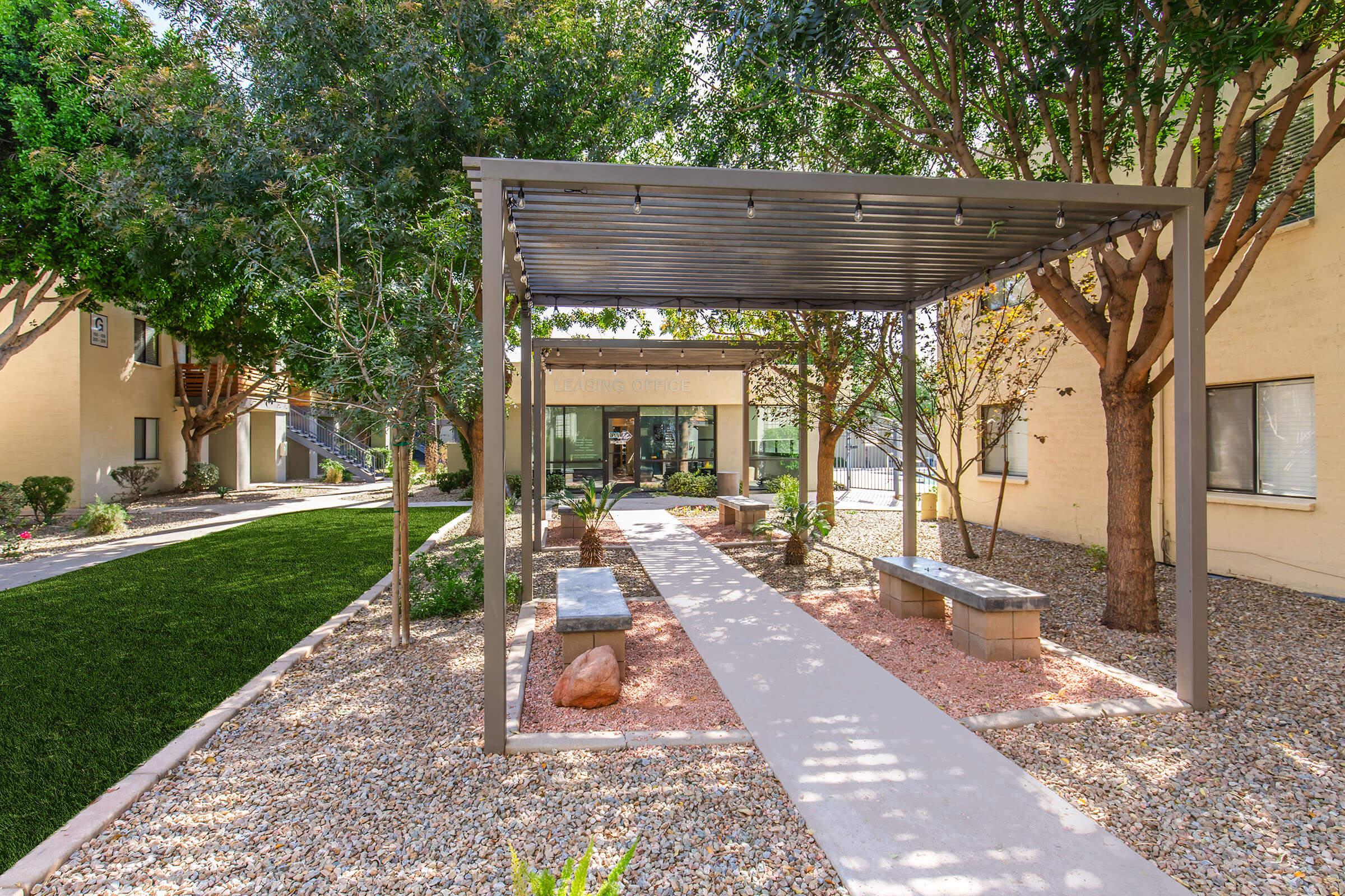 A landscaped courtyard featuring a gravel pathway bordered by trees and benches. A pergola provides shade over the seating area, with green grass and manicured plants enhancing the serene atmosphere. Buildings can be seen in the background, creating a welcoming environment.