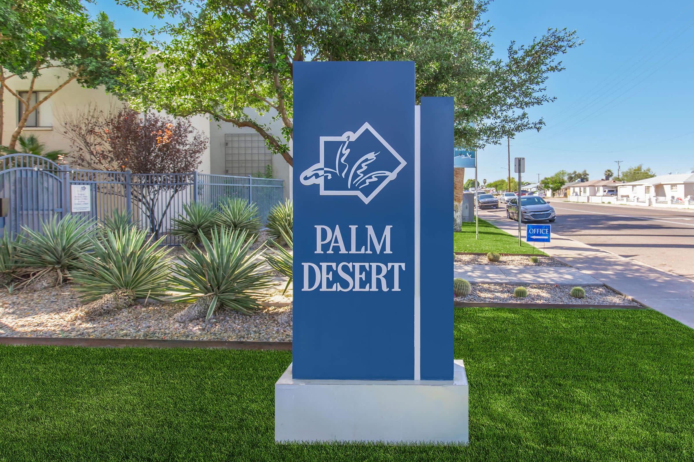 A blue sign displaying the text "PALM DESERT" with a stylized logo featuring palm trees. The sign is positioned on grass near a sidewalk, with shrubs and trees in the background. The setting suggests a welcoming entrance or location in a desert area.