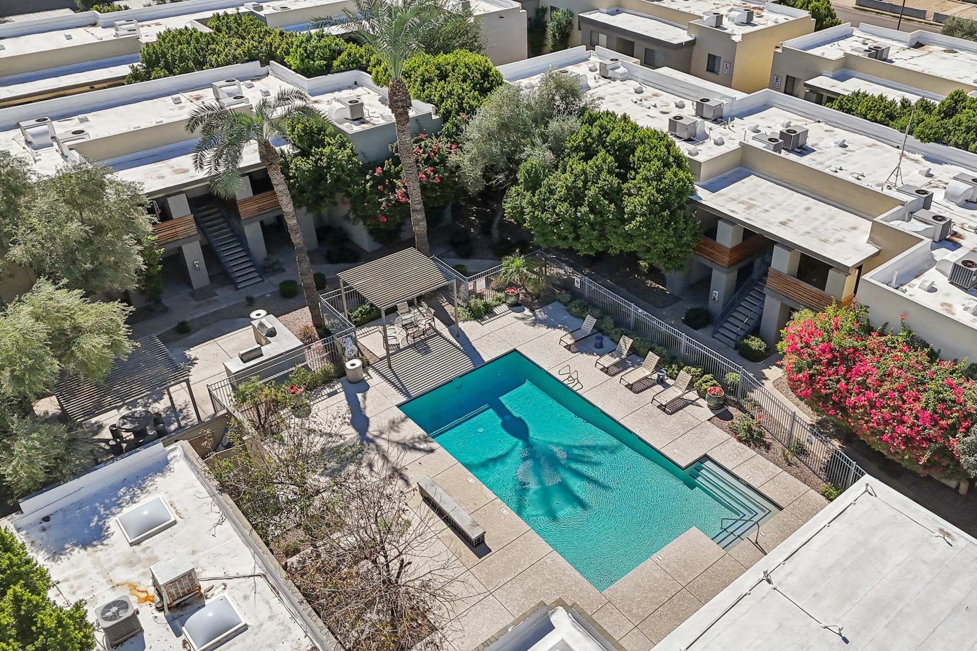 Bird's-eye view of a residential complex featuring a central swimming pool surrounded by lounge chairs and lush greenery. The area is adorned with flowering plants and palm trees, with buildings lining the perimeter, creating a serene and inviting atmosphere.