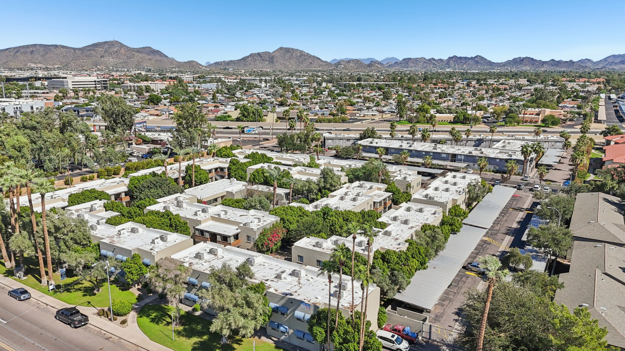 Aerial view of a residential area featuring low-rise apartment buildings surrounded by greenery and trees, with mountains in the background. The scene shows a mix of urban and natural elements, with roads and parked cars visible. Clear blue sky adds to the vibrant atmosphere of the neighborhood.