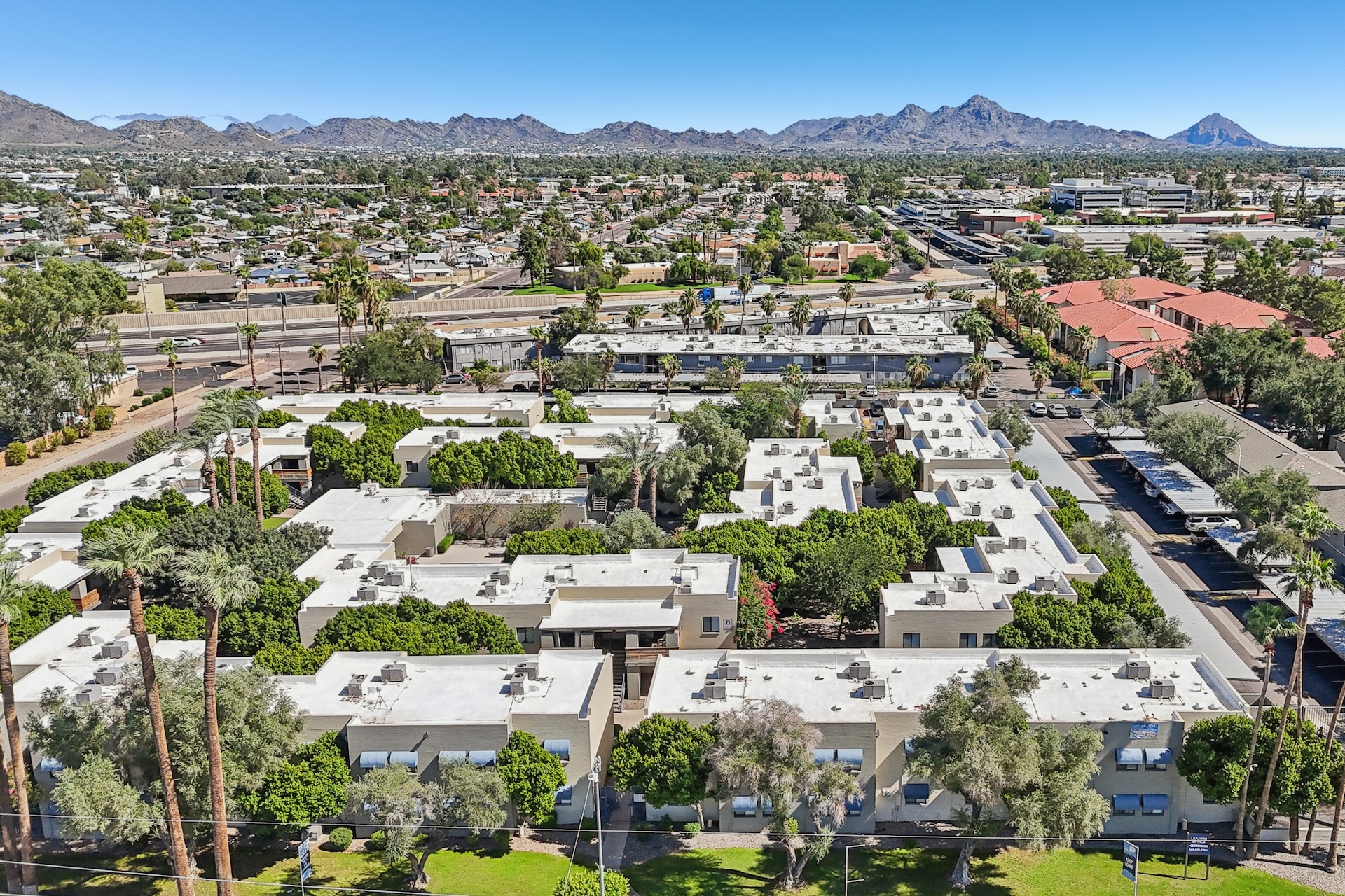 Aerial view of a residential area featuring multiple low-rise buildings surrounded by greenery. The landscape includes mountains in the distance, a clear blue sky, and a highway visible nearby. The scene showcases a mix of urban and natural elements.