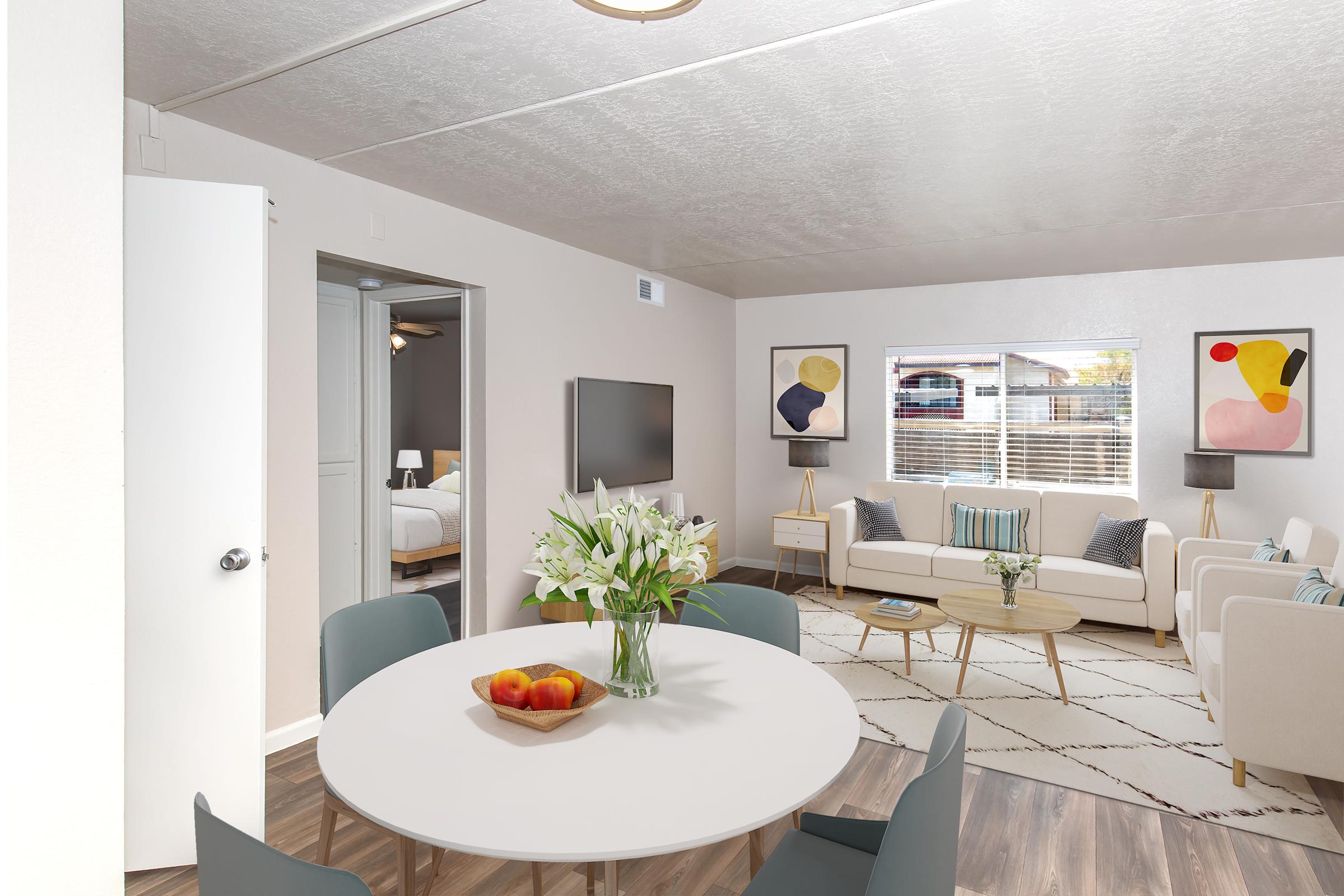 A stylish living area featuring a round dining table with a vase of flowers and a bowl of apples. The room has modern furniture, including a sofa set and a television, with large windows allowing natural light. Abstract art is displayed on the walls, creating a contemporary ambiance.