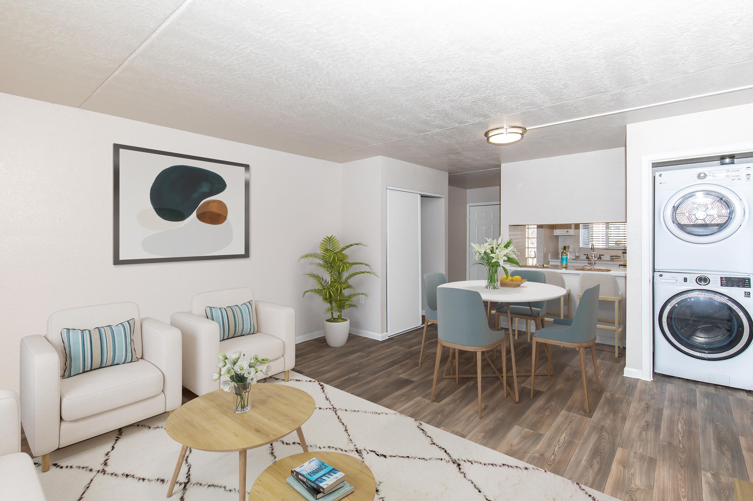 A bright and modern living space featuring two white chairs with blue cushions, a round dining table with four chairs, a potted plant, and a decorative painting on the wall. The kitchen area is visible in the background, alongside a stacked washer and dryer. The flooring is light wood, and there's a cozy rug under the table.