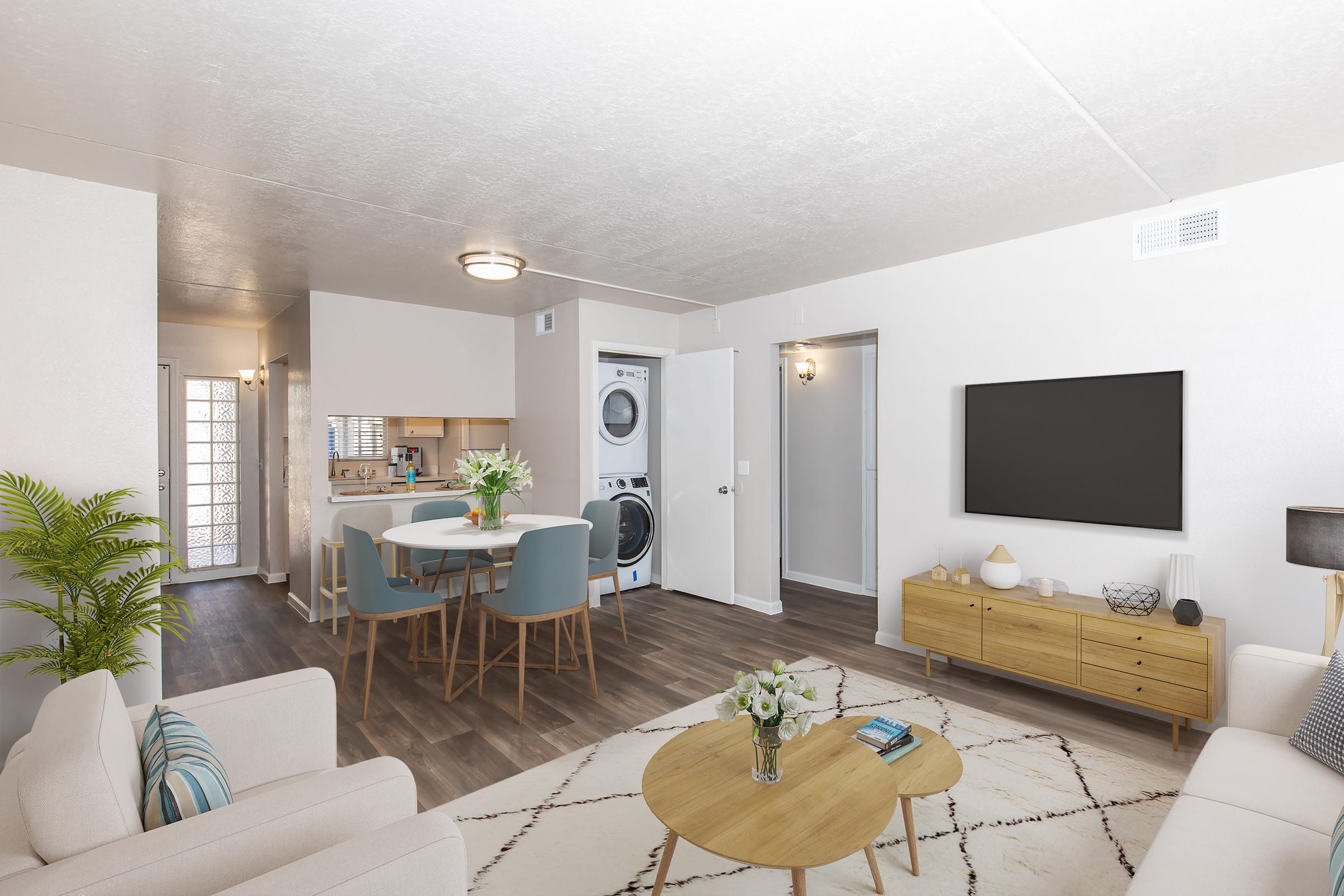 A modern living room with a cozy sofa, round coffee table, and a TV on a wooden stand. Adjacent is a dining area with a round table and blue chairs. In the background, there's a laundry area with stacked washer and dryer. Natural light fills the space, enhancing the neutral color palette and decor.