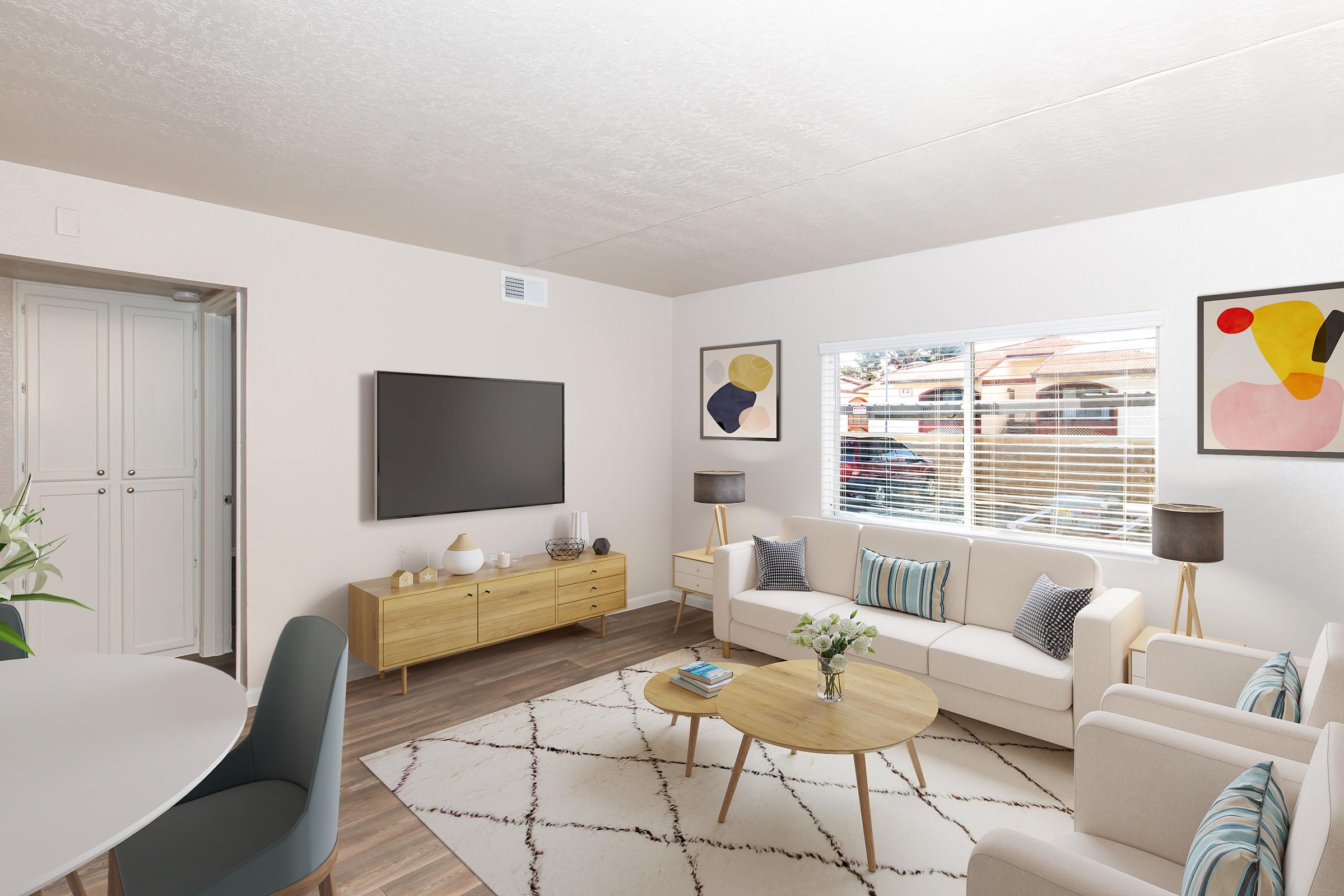 A bright, modern living room featuring a white sofa and a wooden coffee table on a patterned area rug. There's a TV mounted on the wall, and two lamps beside the sofa. Abstract artwork hangs on the walls, and a dining area with blue chairs is visible in the background, along with a window letting in natural light.