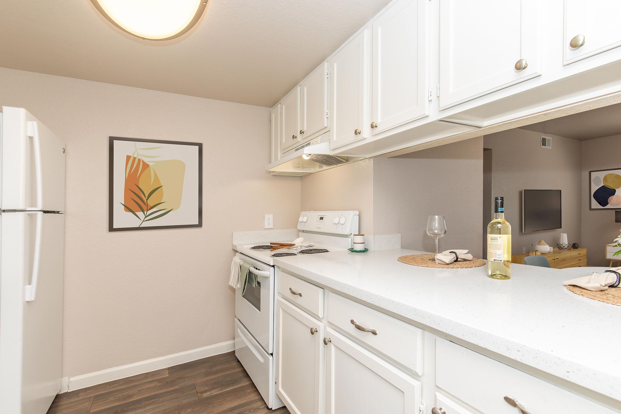 A modern kitchen featuring white cabinetry, a white stove, and a refrigerator. Countertops are adorned with a bottle of white wine and a glass. A piece of framed wall art displaying abstract shapes hangs nearby. The open layout allows a glimpse into a living area with additional decor.