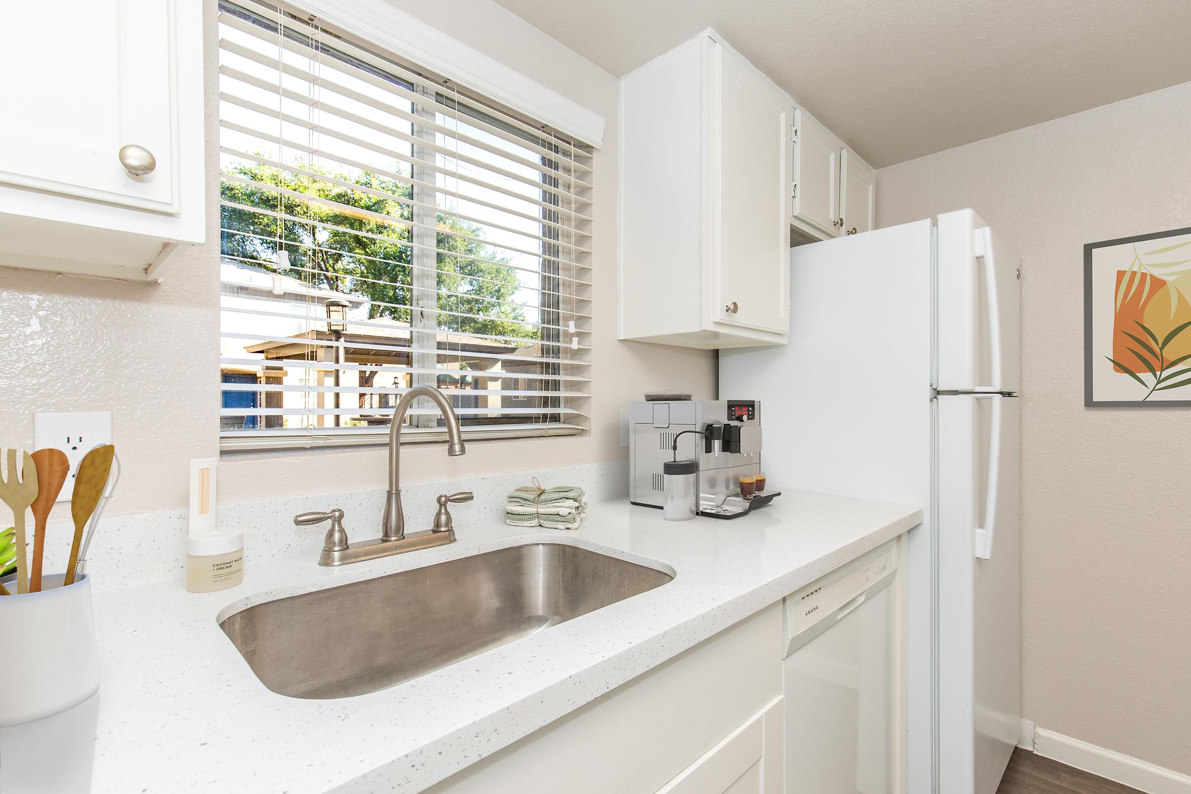A bright and modern kitchen featuring a stainless steel sink, white cabinets, and a refrigerator. Natural light comes in through a window, showcasing a small outdoor area. On the countertop, there are kitchen utensils, a soap dispenser, and a coffee maker, adding to the space's functionality.