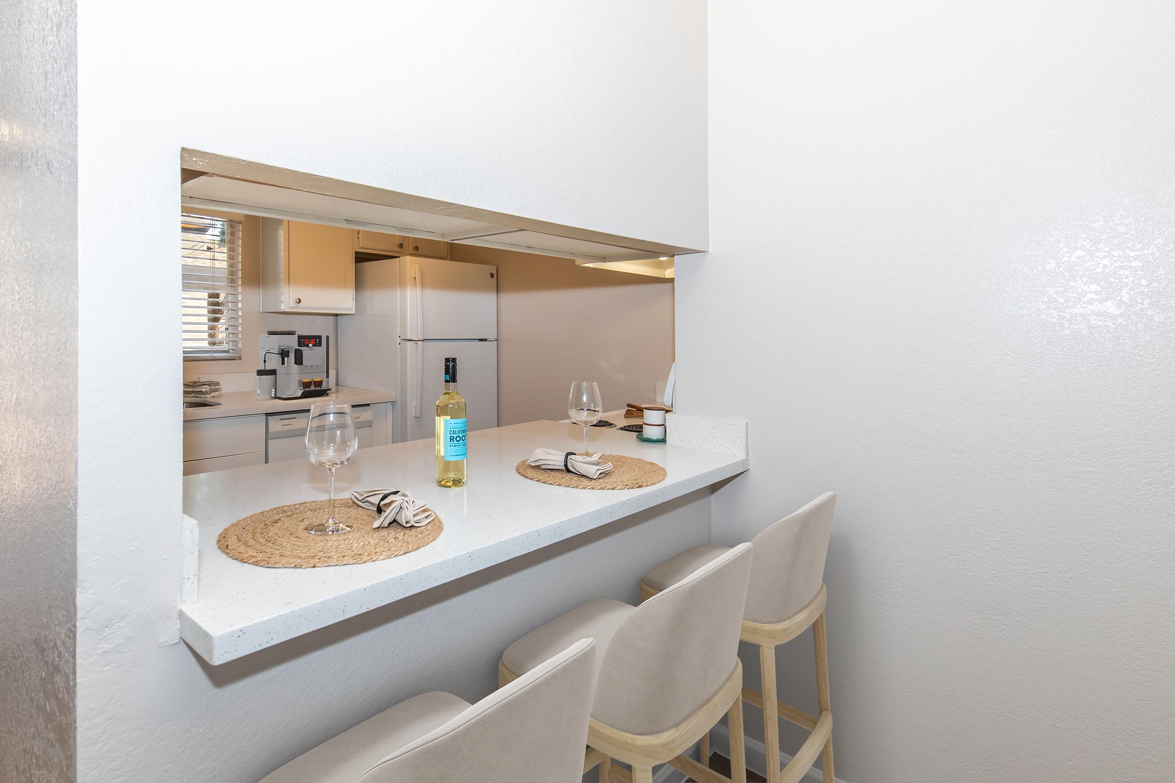 A modern kitchen featuring a pass-through bar with two beige stools. On the countertop, there are two wine glasses and a bottle of white wine. The kitchen is bright and airy, with white cabinetry and appliances visible in the background. Sunlight streams in through a window, creating a warm atmosphere.
