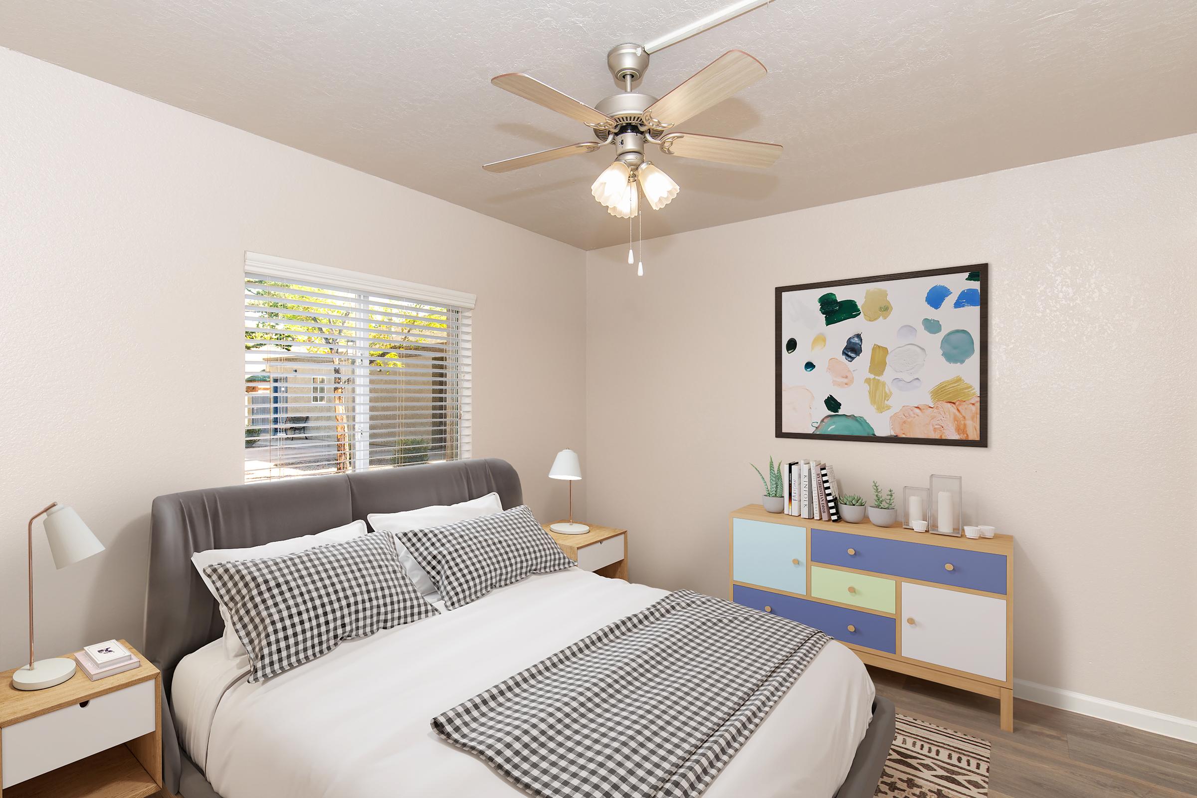 A cozy bedroom featuring a large bed with gray and white bedding, a minimalist wooden nightstand, and a colorful abstract painting on the wall. A stylish dresser with blue and white drawers sits beside the bed, and a ceiling fan provides air circulation. Natural light enters through a window adorned with blinds.