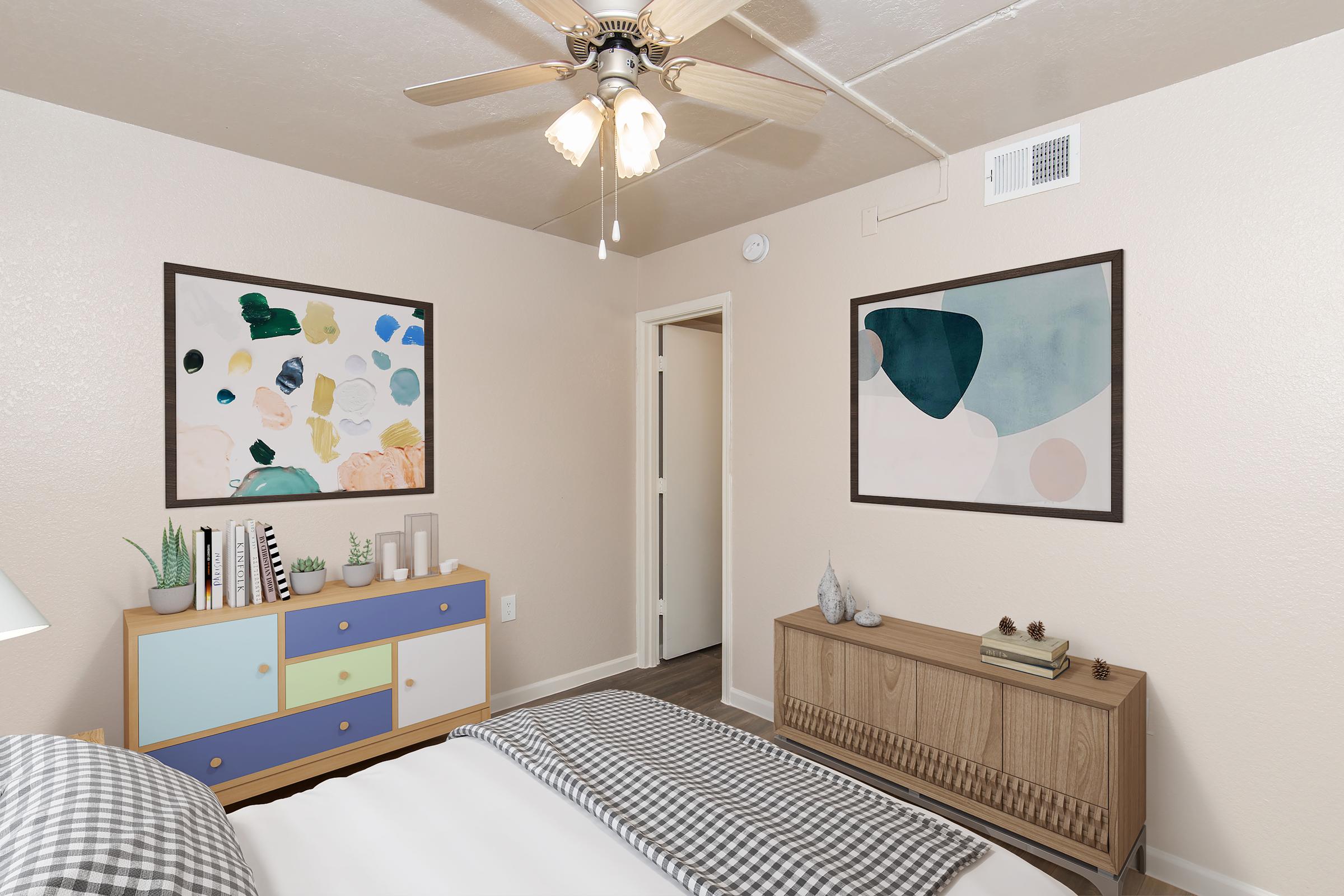 A cozy bedroom featuring a bed with a checkered blanket, a light blue and wood dresser with decorative items, and two framed artworks on the walls. A ceiling fan provides a warm ambiance, and a door leads to another room.