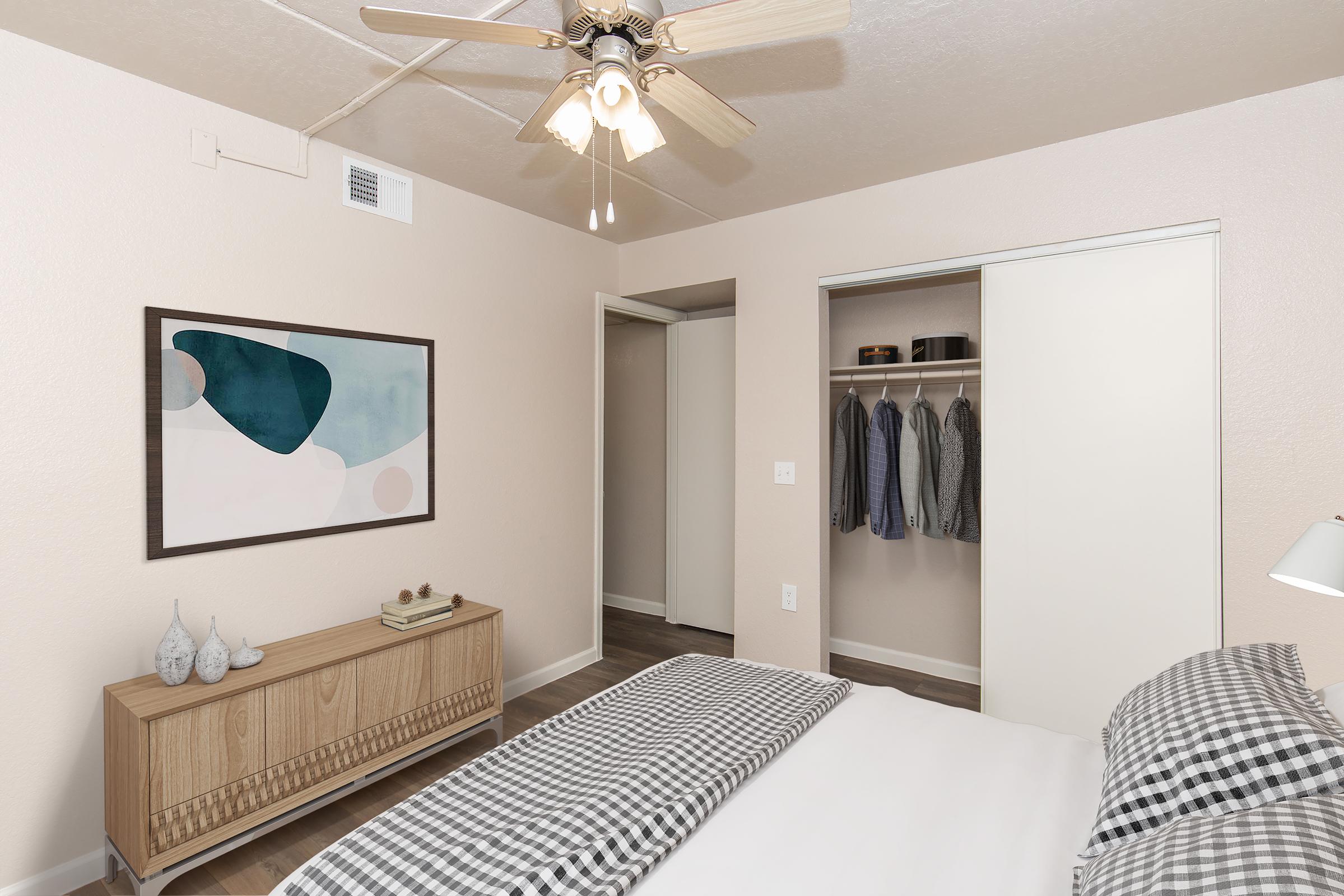 A cozy bedroom featuring a queen-sized bed with a checkered blanket, a wooden dresser with decorative items, and a framed abstract art piece on the wall. A ceiling fan circulates air, and there is a closet with neatly hung clothes visible. The walls are painted a soft beige color, creating a warm ambiance.