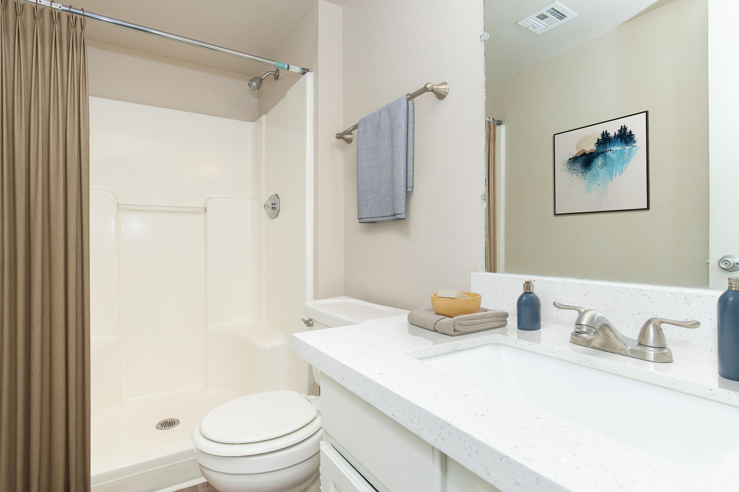 A clean, well-lit bathroom featuring a white shower stall, beige shower curtain, and a modern sink with a speckled countertop. A towel hangs on the wall, and decorative items include a small bowl and a blue vase. A mirror reflects the space, enhancing the room's brightness.