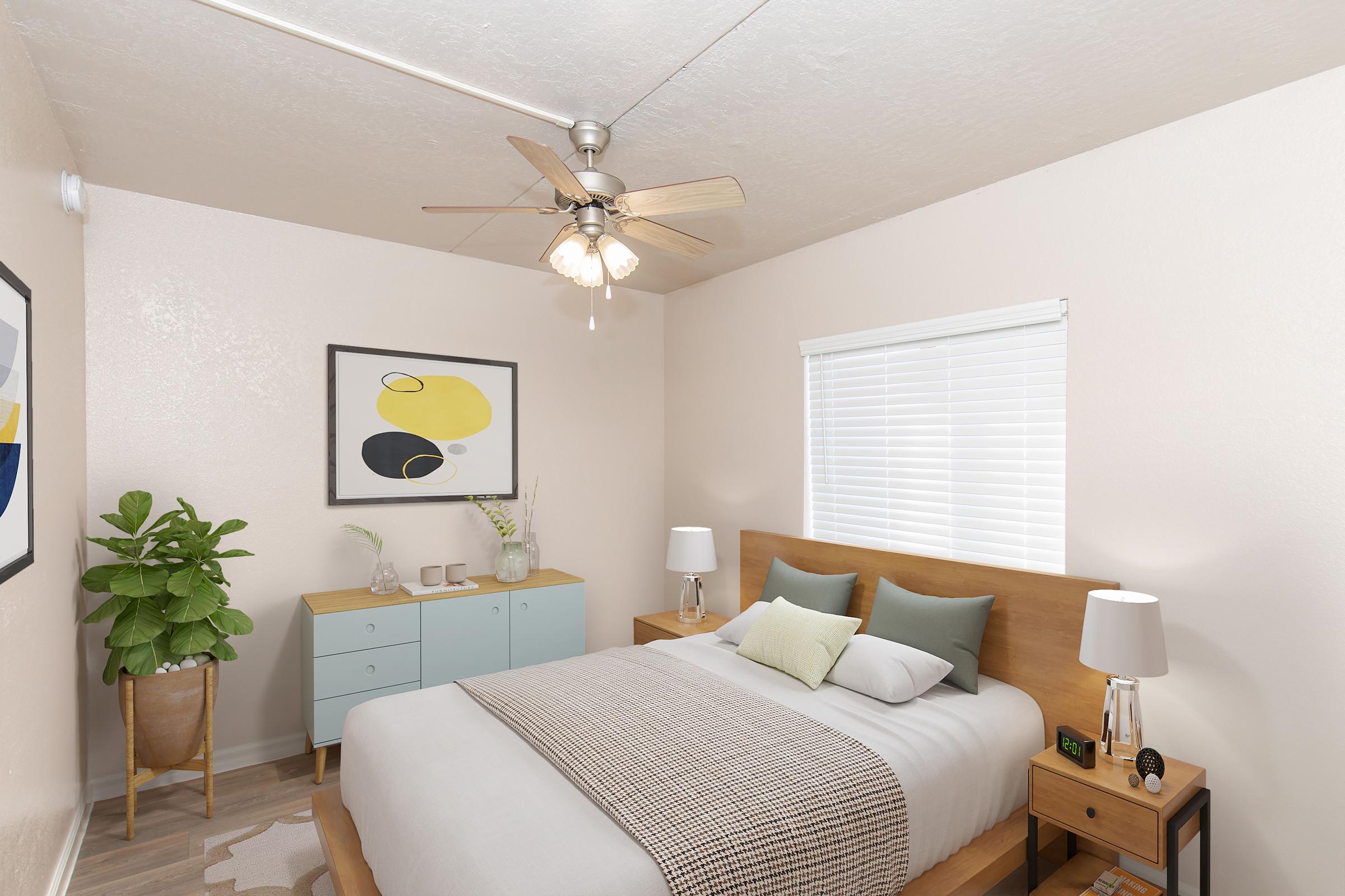 A softly lit bedroom featuring a queen-sized bed with light gray bedding and decorative pillows. There are two bedside lamps on matching nightstands. A light blue dresser is against the wall, decorated with plants and a framed artwork. A ceiling fan is overhead, and a window with white blinds allows natural light to enter.