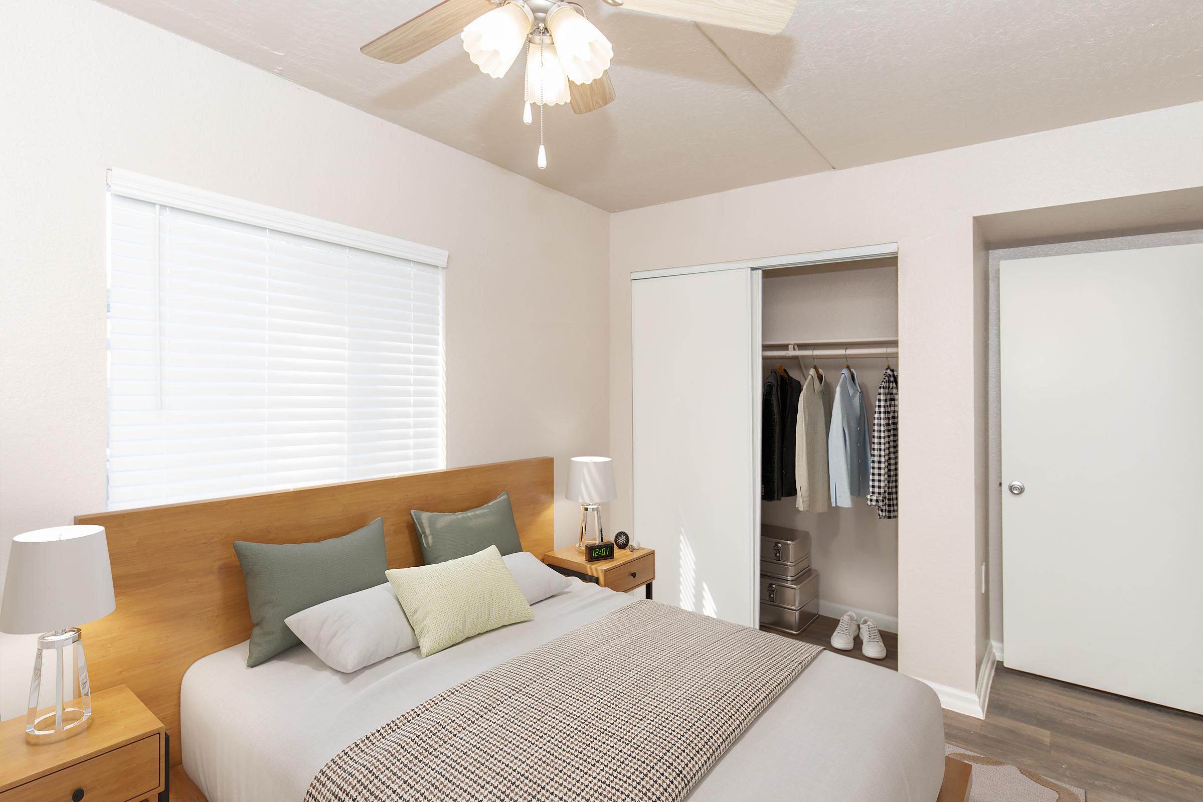 A cozy bedroom featuring a wooden bed with decorative pillows, two bedside lamps, and a patterned throw. There's a closet with sliding doors, showcasing neatly hung clothes and shoes. Natural light filters through a window with white blinds, enhancing the serene atmosphere of the room.