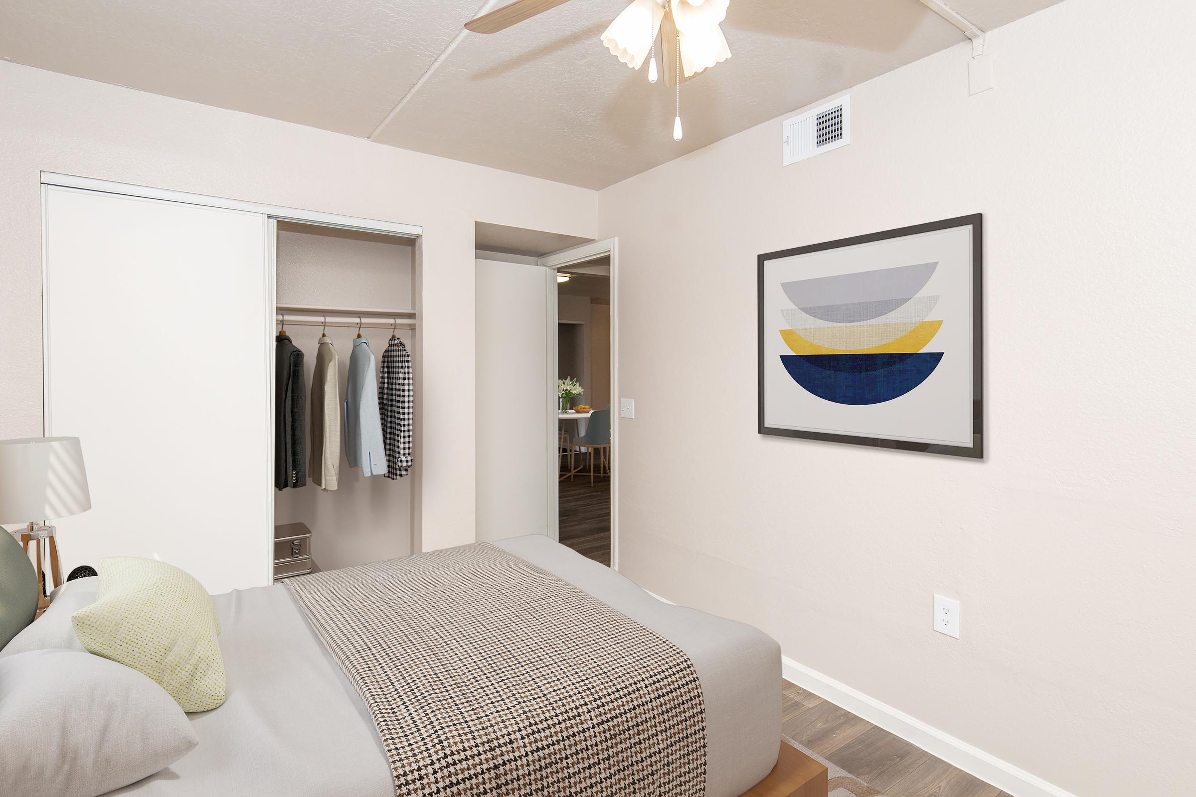 A cozy bedroom featuring a neatly made bed with a gray blanket and decorative pillows. There is a framed abstract art piece on the wall, and a closet with hanging clothing. A ceiling fan is visible, and part of a living space with a dining area can be seen through the open door in the background.