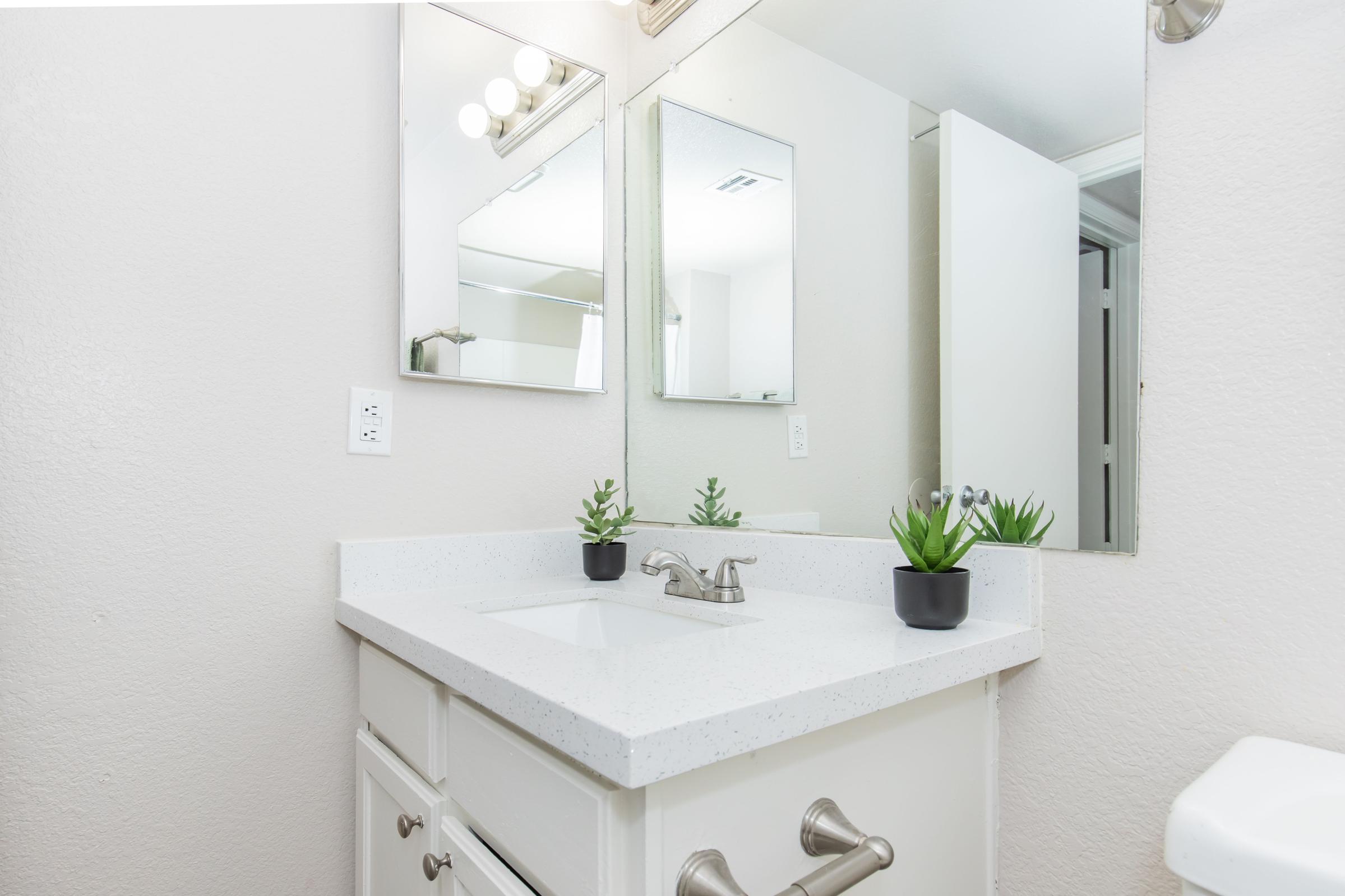 A well-lit bathroom featuring a white countertop with a sink, two mirrors above it, and a potted succulent on the countertop. The walls are painted light, and a partially opened door leads to another room, enhancing the spacious feel of the area.
