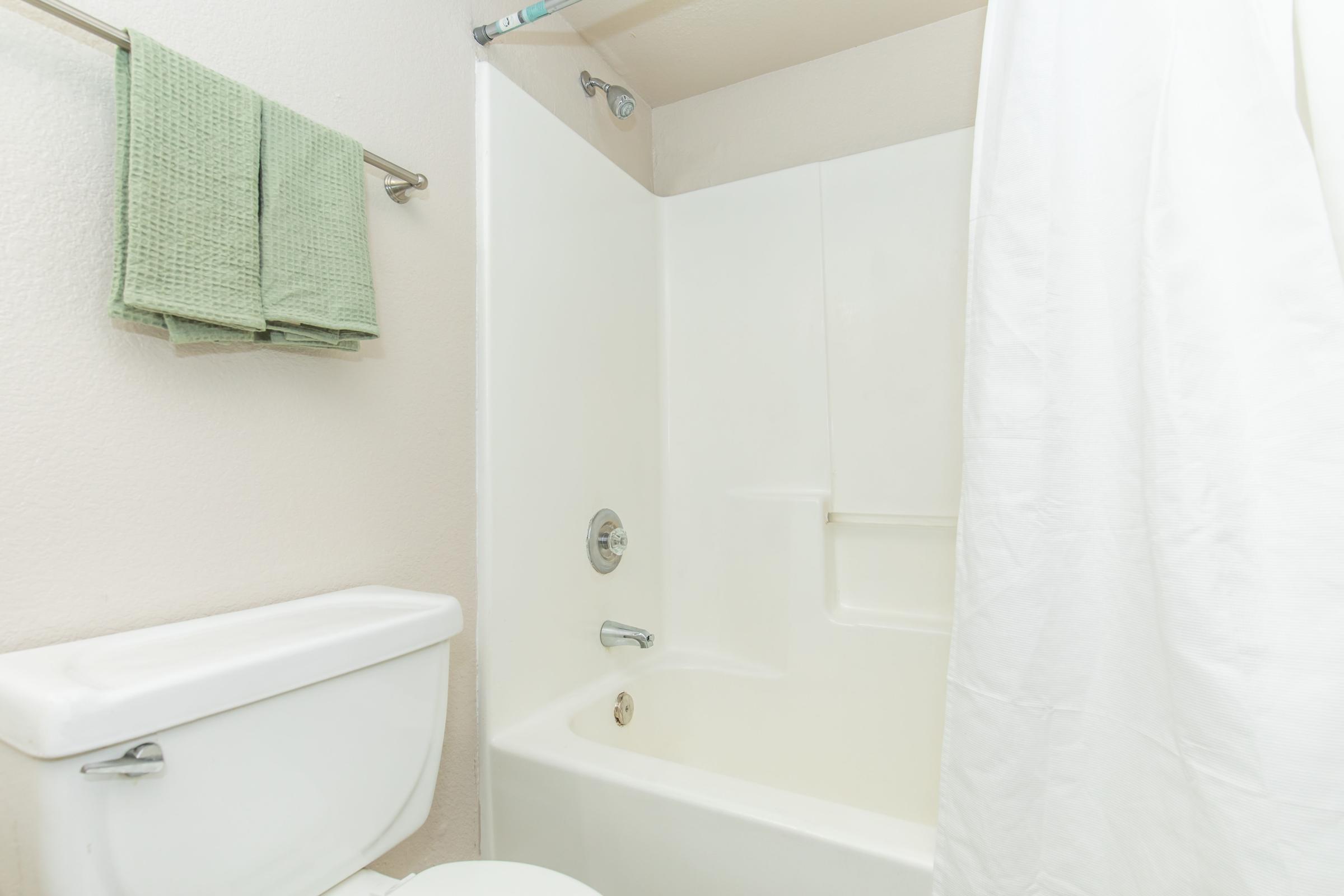 A clean bathroom featuring a white bathtub and shower. There is a white shower curtain and green towels hanging on a towel rack. The toilet is visible in the foreground, and the walls are a light color, creating a bright and fresh appearance.