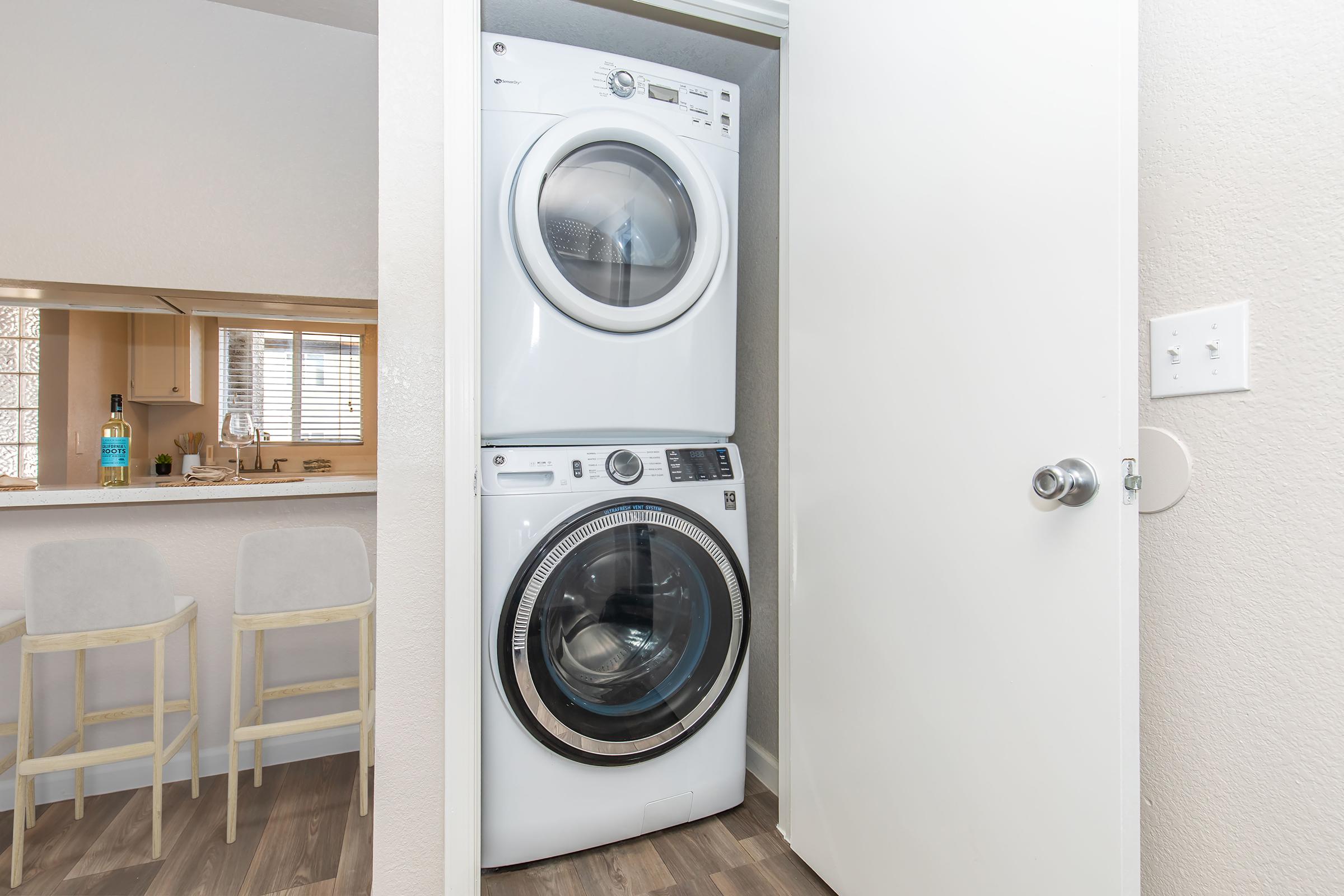 A compact laundry area featuring a stacked washer and dryer in a small closet. The space shows light-colored walls and hardwood flooring, with glimpses of a bar-style seating area in the background.
