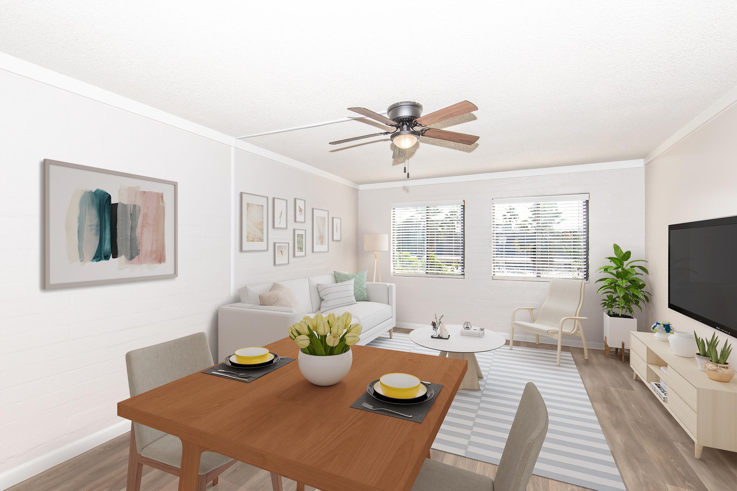 A bright and airy living space featuring a dining area with a wooden table set for two, a cozy couch, and a coffee table. The room includes large windows with blinds, a wall-mounted TV, a ceiling fan, and decorative art on the walls. A green plant adds a touch of nature to the modern decor.