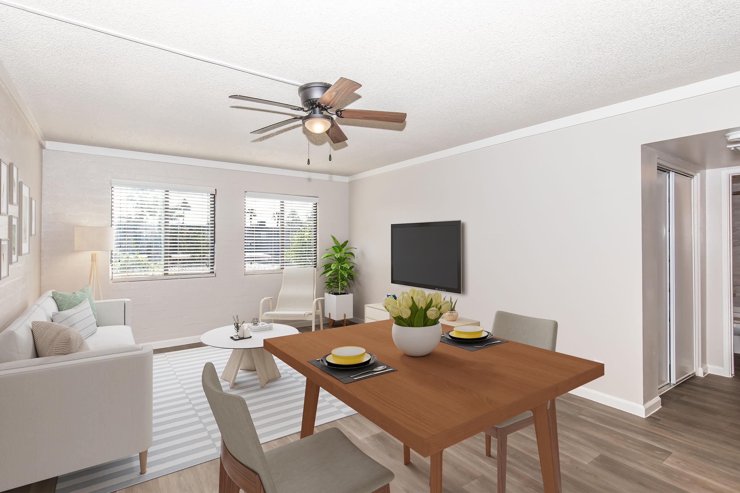 Bright and airy living space featuring a comfortable seating area with a light-colored couch, a coffee table, and two chairs. A dining table with place settings is visible, along with a ceiling fan and a television mounted on a white wall. Natural light floods in through windows, and a potted plant adds a touch of greenery.