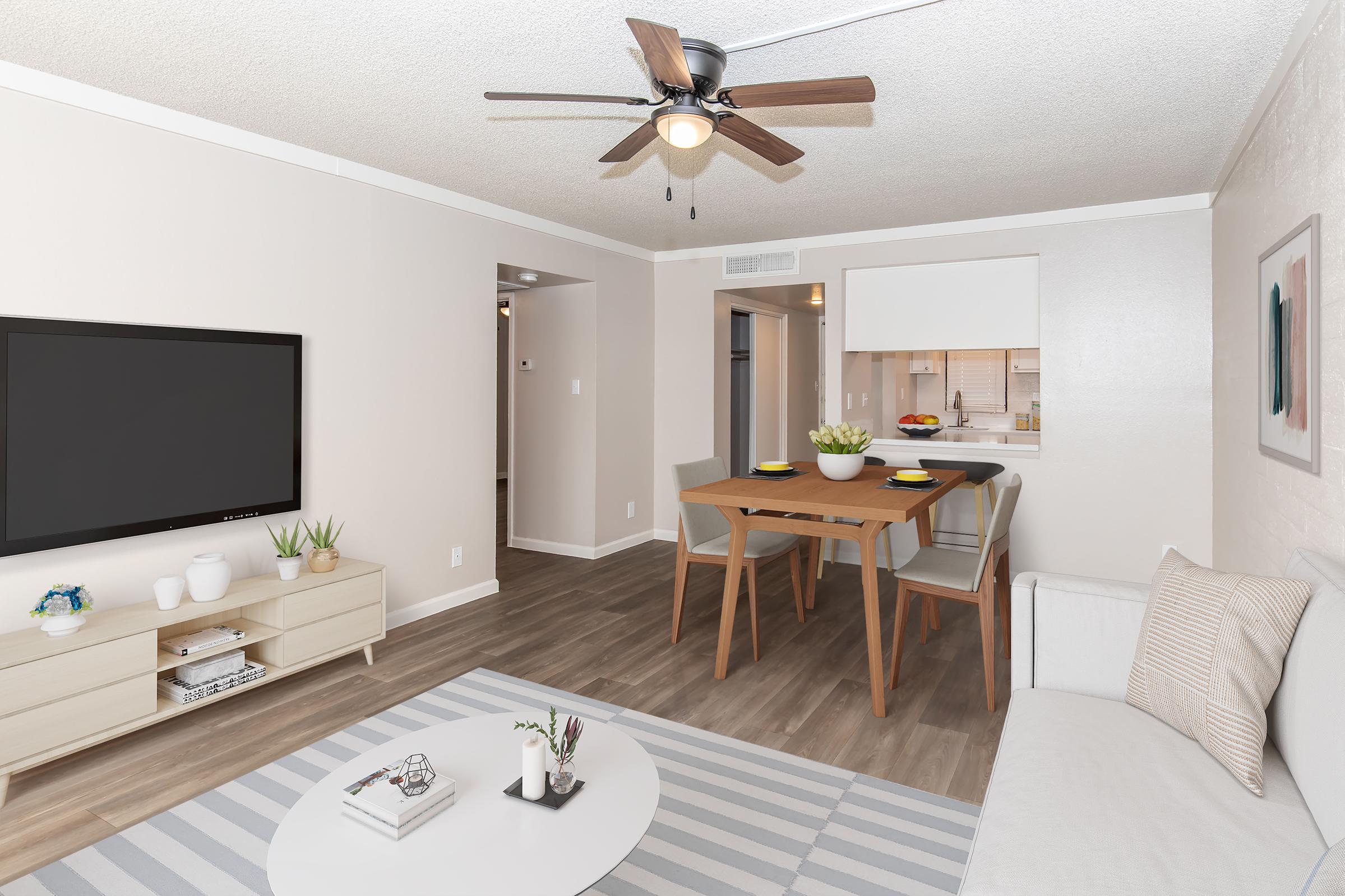 A modern living room featuring a light-colored sofa, a round coffee table, and a television mounted on the wall. A dining area with a wooden table and two chairs is visible, along with a well-lit kitchen in the background. Neutral-colored walls and wooden flooring create a cozy atmosphere.