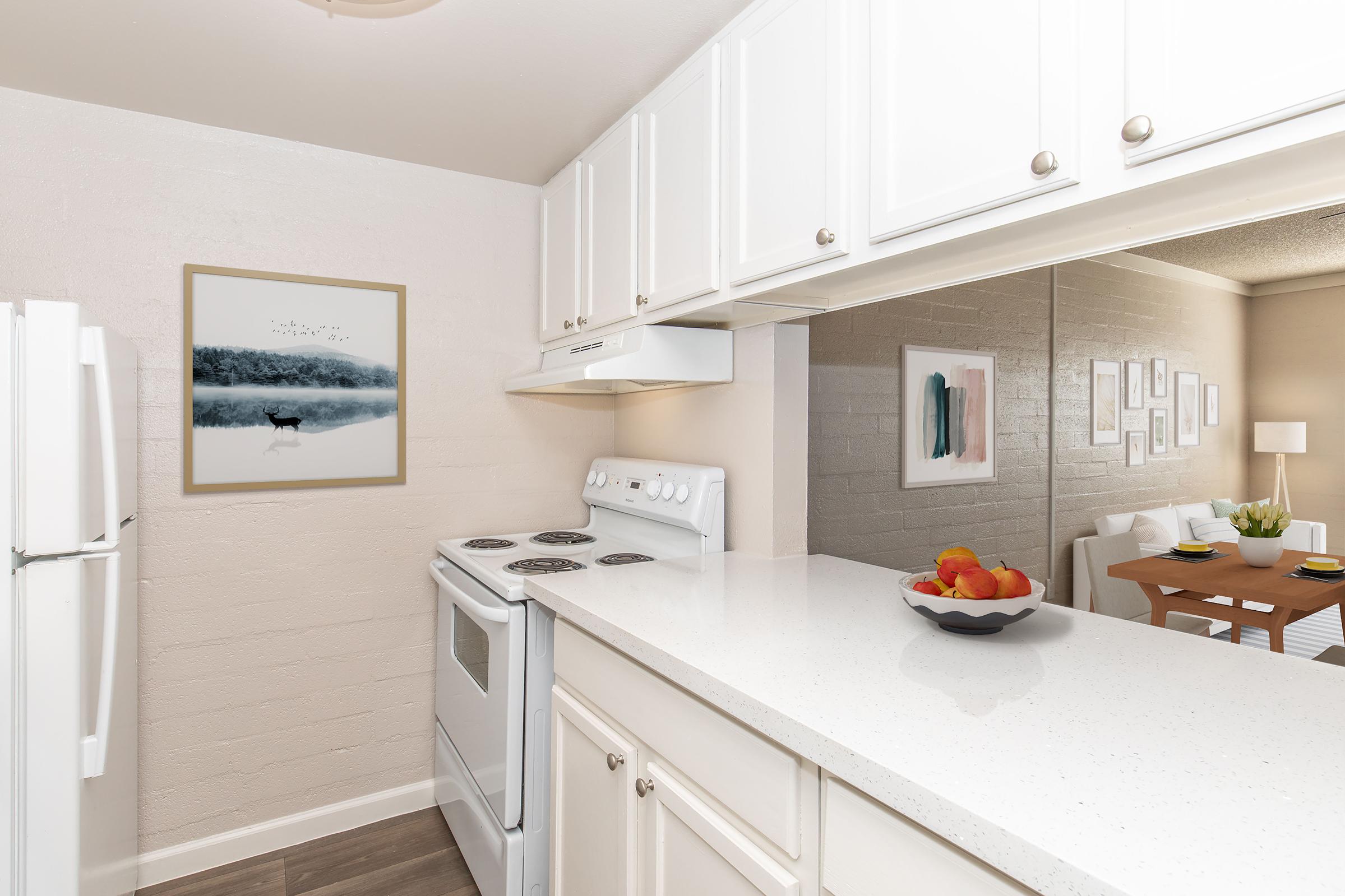 A bright and modern kitchen featuring white cabinetry, a stove, and a countertop with a bowl of colorful fruit. A wall-mounted art piece is displayed, showing a tranquil landscape. Reflections in the mirrored wall add depth to the space, and a dining area is visible in the background with a table set for meals.