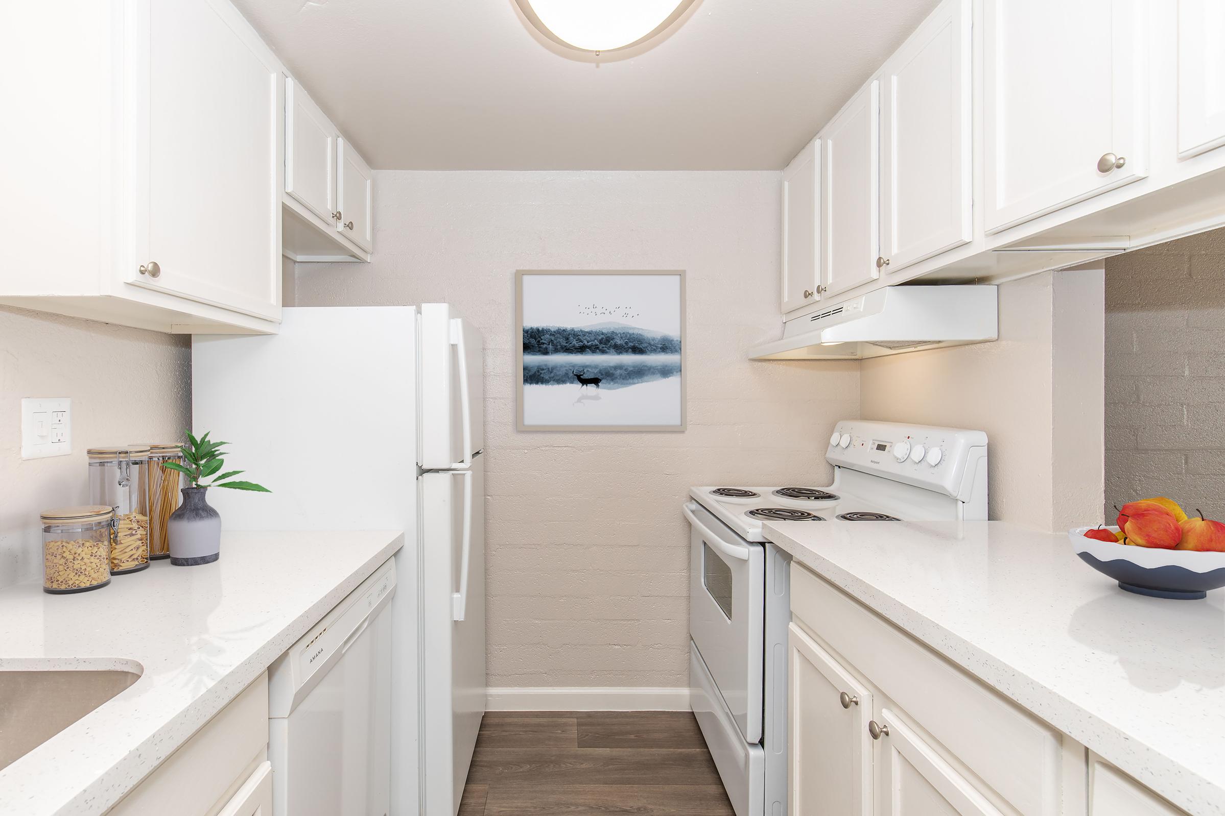 A modern kitchen featuring white cabinets, a refrigerator, and an oven. The countertop is light-colored with a bowl of apples and a plant. A framed picture of a serene landscape hangs on the wall, completing the tidy and stylish look of the space.