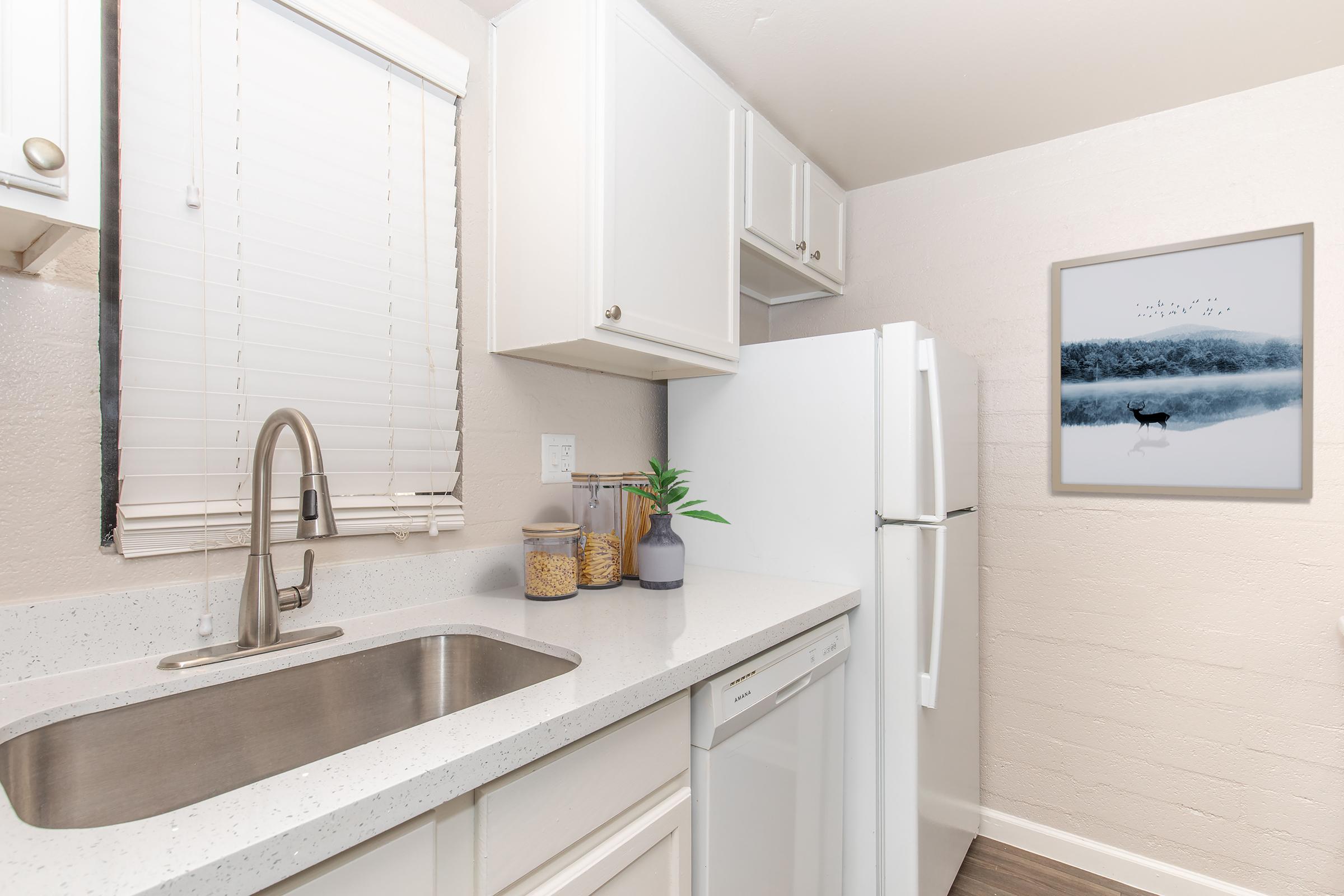 Modern kitchen with white cabinetry, stainless steel sink, and countertop. A refrigerator and dishwasher are visible. A jar of grains and a potted plant sit by the sink. A framed artwork depicting a serene landscape with a deer hangs on the wall. Soft natural light enters through a window with blinds.