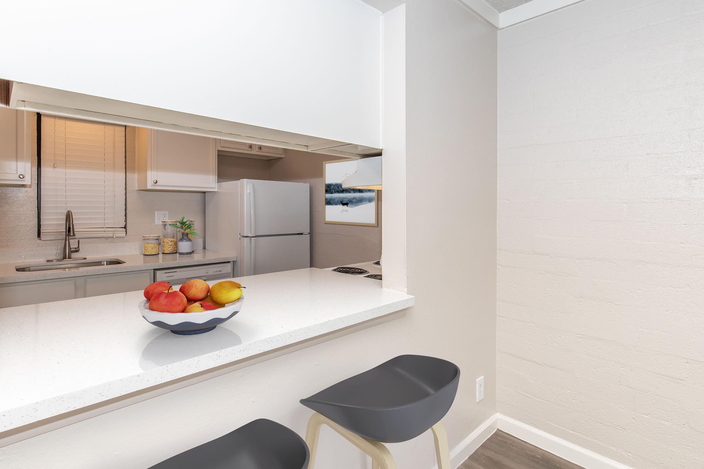 A modern kitchen with a white countertop and bar seating. A bowl of colorful fruits sits on the counter. In the background, there’s a white refrigerator, a sink, and a window with natural light. The overall design is clean and minimalist, featuring light-colored walls and wooden flooring.