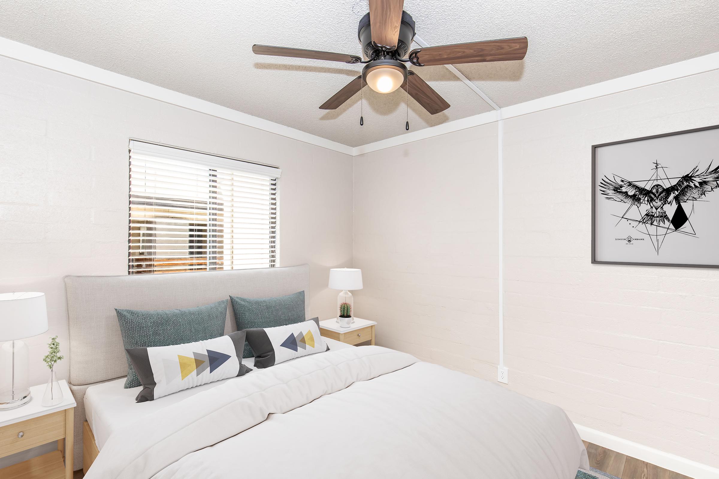 A cozy bedroom featuring a light-colored wall, a queen-sized bed with a beige headboard and decorative pillows in geometric patterns. There's a ceiling fan, wooden nightstands with lamps, and a framed artwork on the wall depicting an eagle. Natural light streams through the window with blinds.