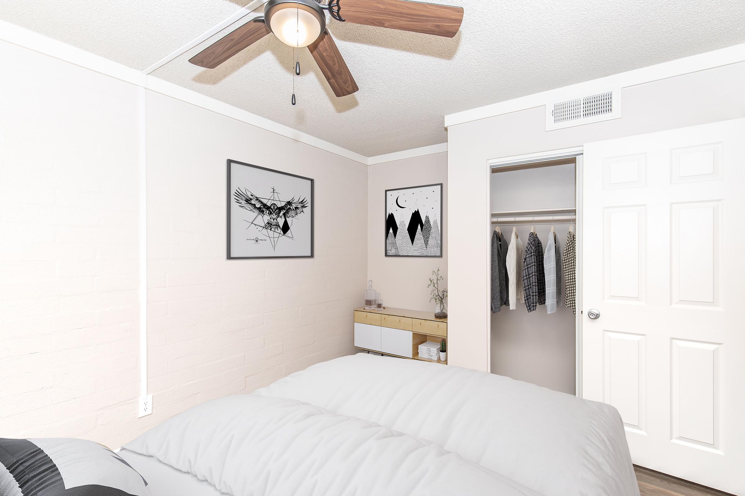 A cozy bedroom featuring a ceiling fan, a neatly made bed with gray bedding, and two framed artworks on the wall. A closet with hanging clothes is visible next to a light wood dresser. Natural light brightens the space, giving it a warm and inviting atmosphere.