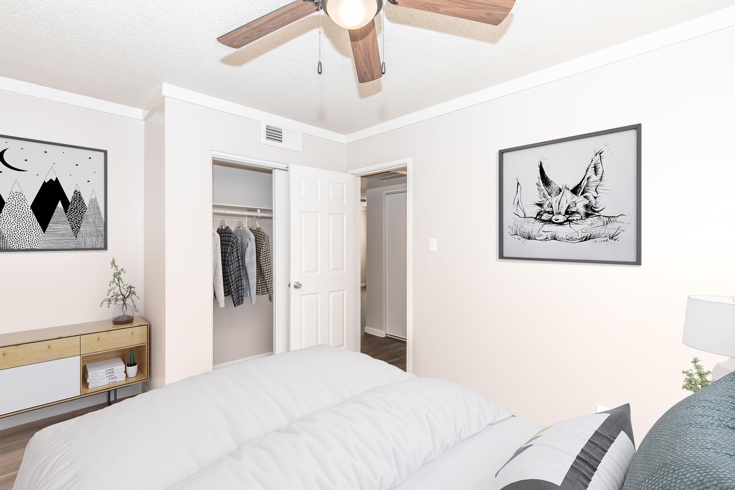 A cozy bedroom featuring a light-colored wall, a ceiling fan, a framed monochrome artwork, and a small nightstand. A closet with clothes is visible through an open door. The bed has a white comforter, creating a welcoming atmosphere.