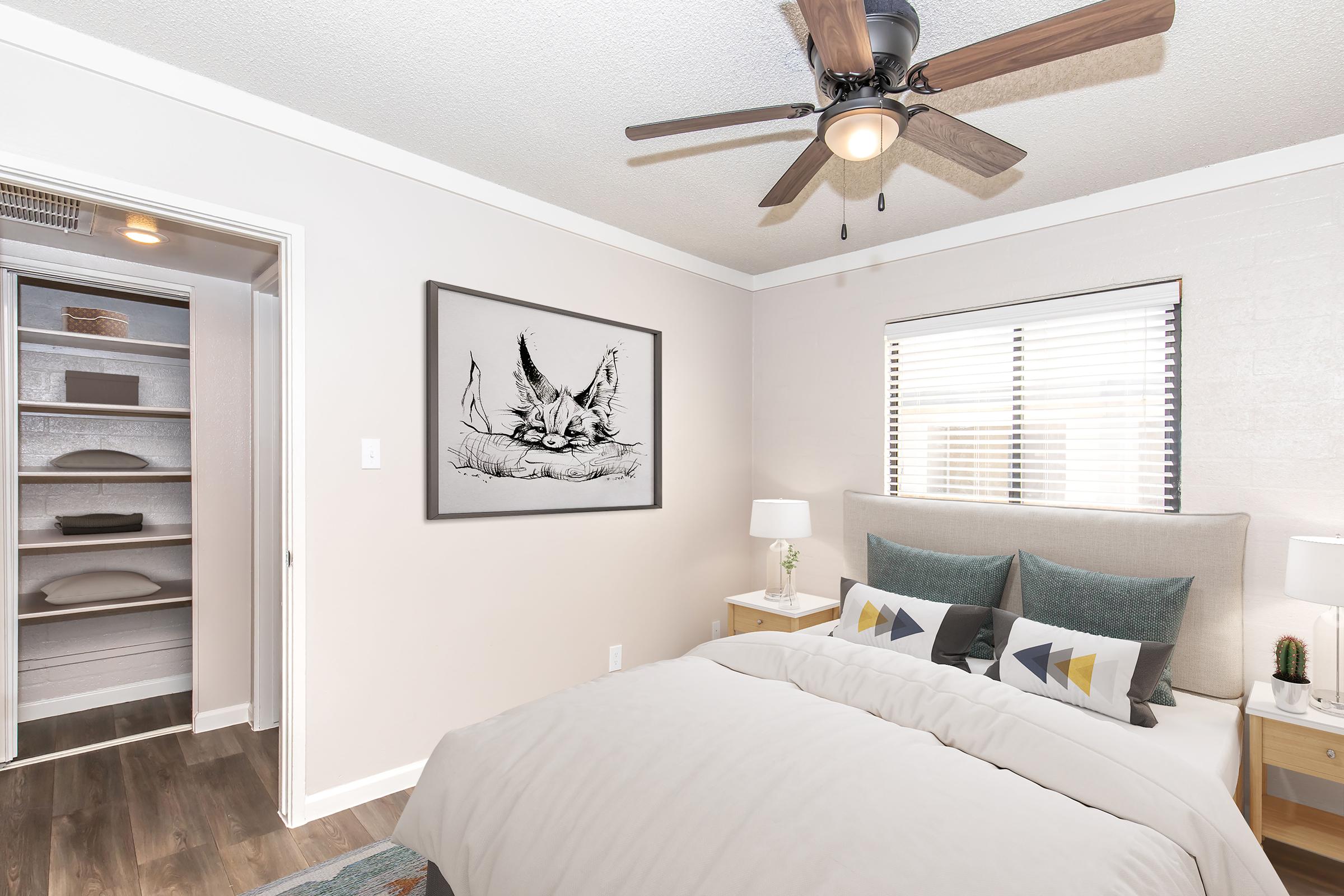 A modern bedroom featuring a light color scheme with neutral walls, a cozy bed adorned with decorative pillows, and a stylish piece of artwork hanging above it. There are bedside tables with lamps, and a ceiling fan above. A closet with shelves is visible through an open doorway. Natural light enters through the window.