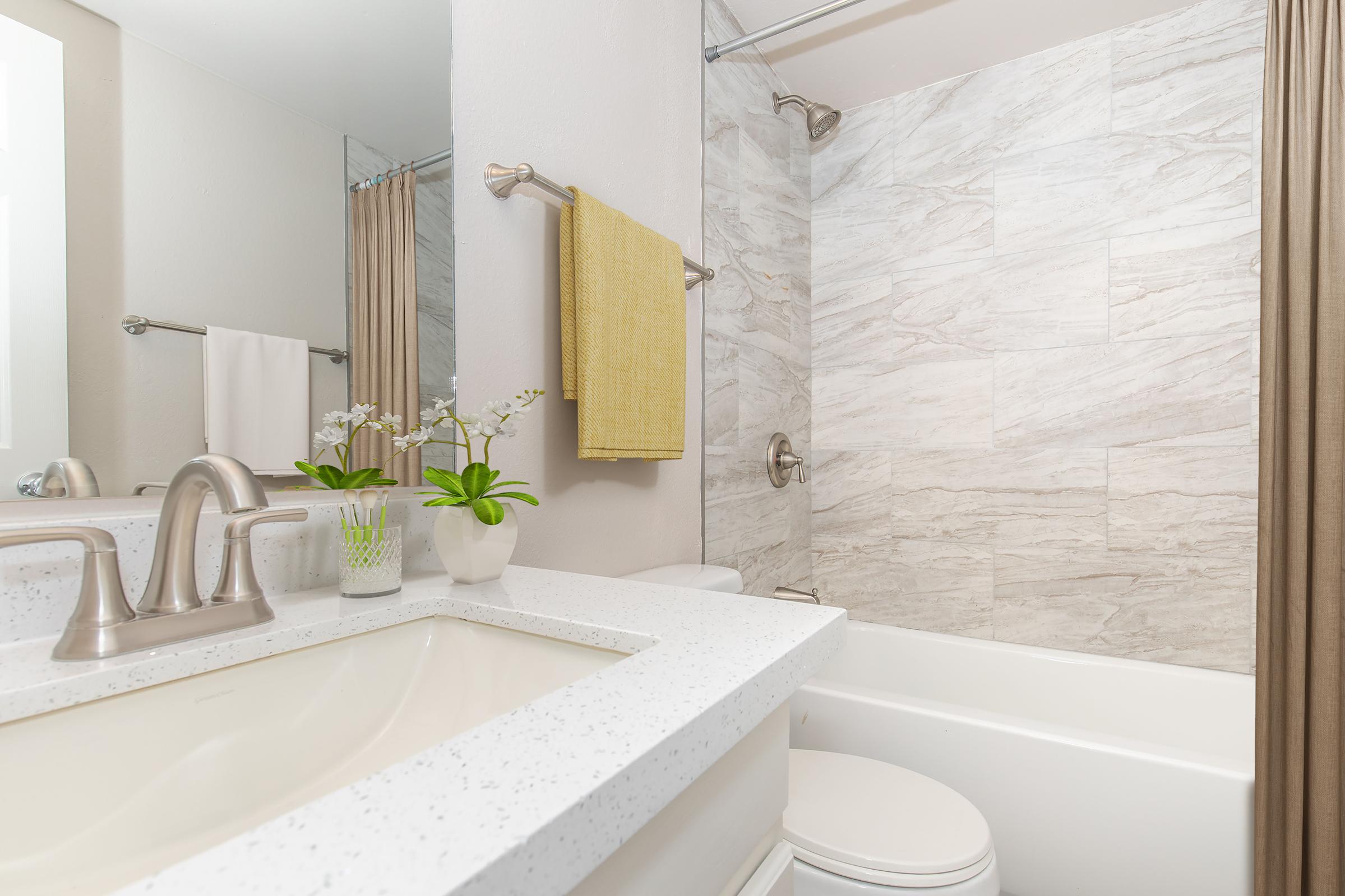 A modern bathroom featuring a white countertop with a sink, a bathtub, and a shower. The walls are tiled in light grey stone, and there's a plant and decorative items on the counter. A yellow towel hangs nearby, adding a pop of color to the neutral palette. The space is clean and well-lit.