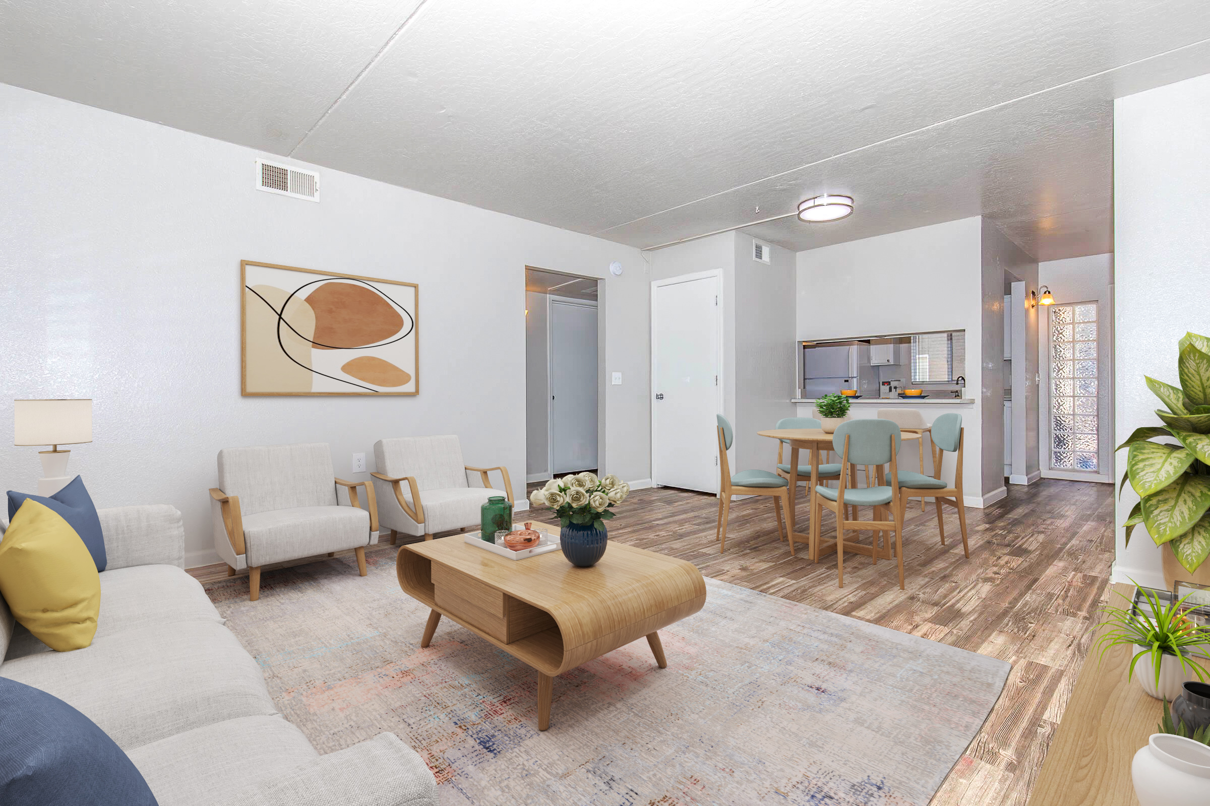 A modern living room with light-colored walls and wooden flooring. It features a comfortable seating area with two armchairs and a sofa, a round coffee table, and a decorative rug. A dining area with a table and chairs is visible in the background, along with a small kitchen. The space is neatly decorated with plants and artwork.