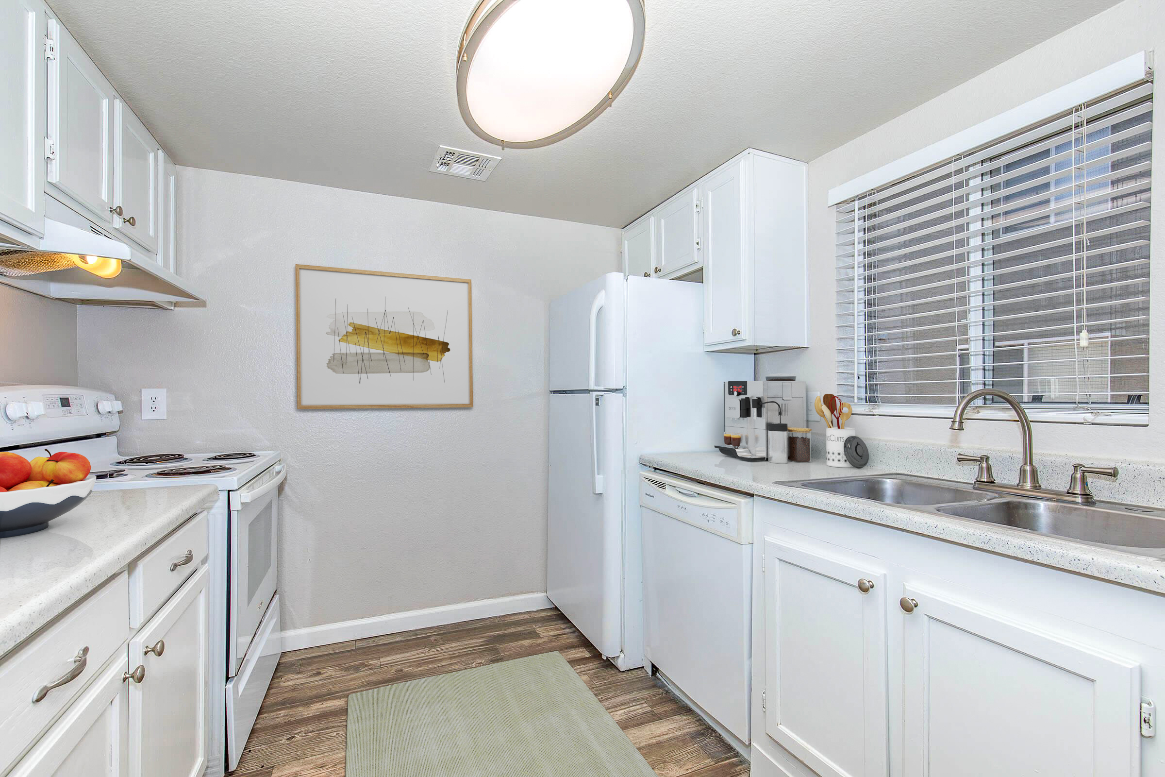 A modern kitchen featuring white cabinets, a refrigerator, an oven, and a sink. There is a small window with blinds allowing natural light, and a framed abstract art piece on the wall. A light-colored rug is on the floor, and some apples are placed on the countertop.