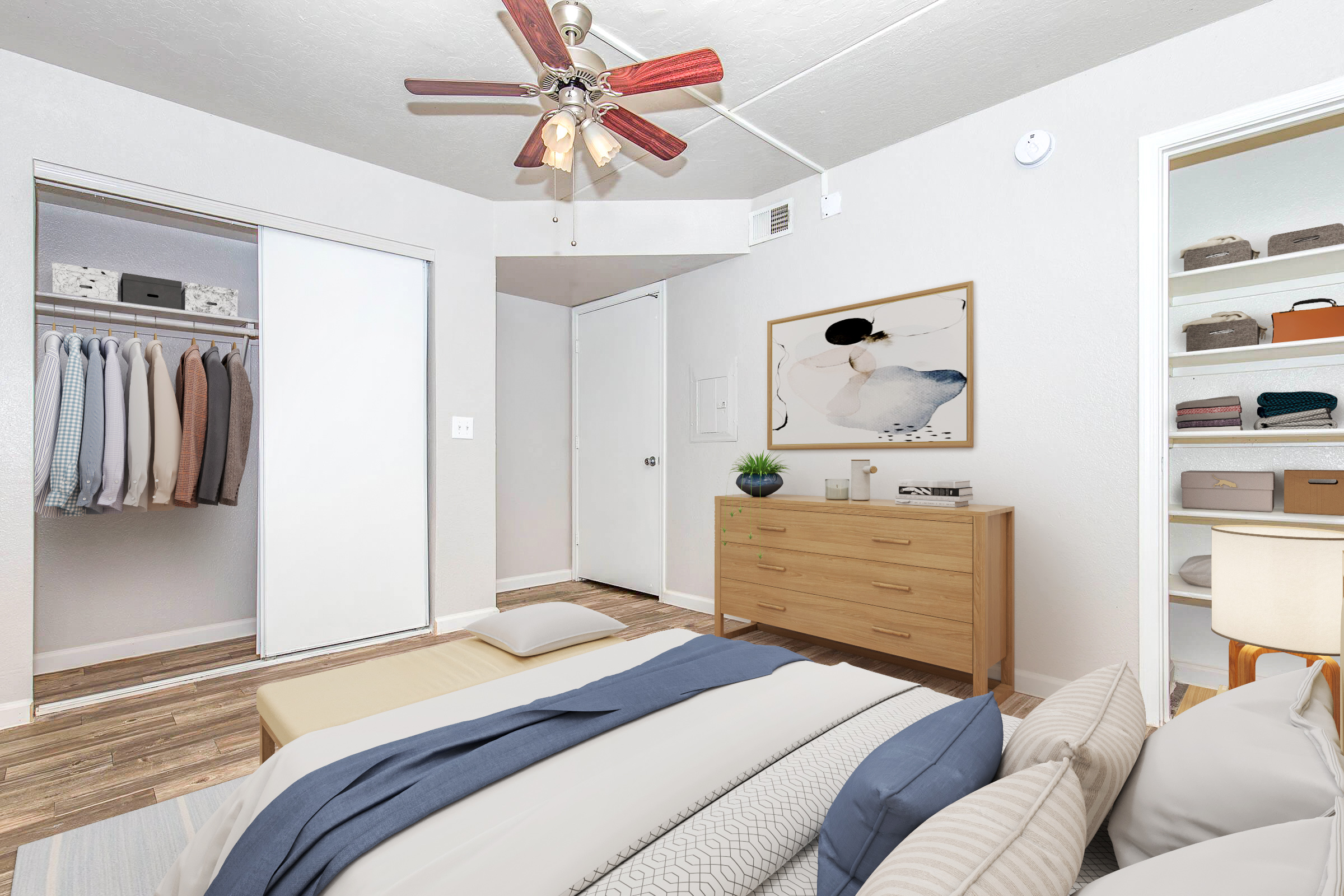 A cozy bedroom featuring a bed with blue and gray bedding, a wooden dresser, and a ceiling fan. A closet with neatly hung shirts is visible on the left, while a white door leads to another room. A piece of abstract art hangs on the wall, and the floor is made of wood. Soft lighting creates a welcoming atmosphere.