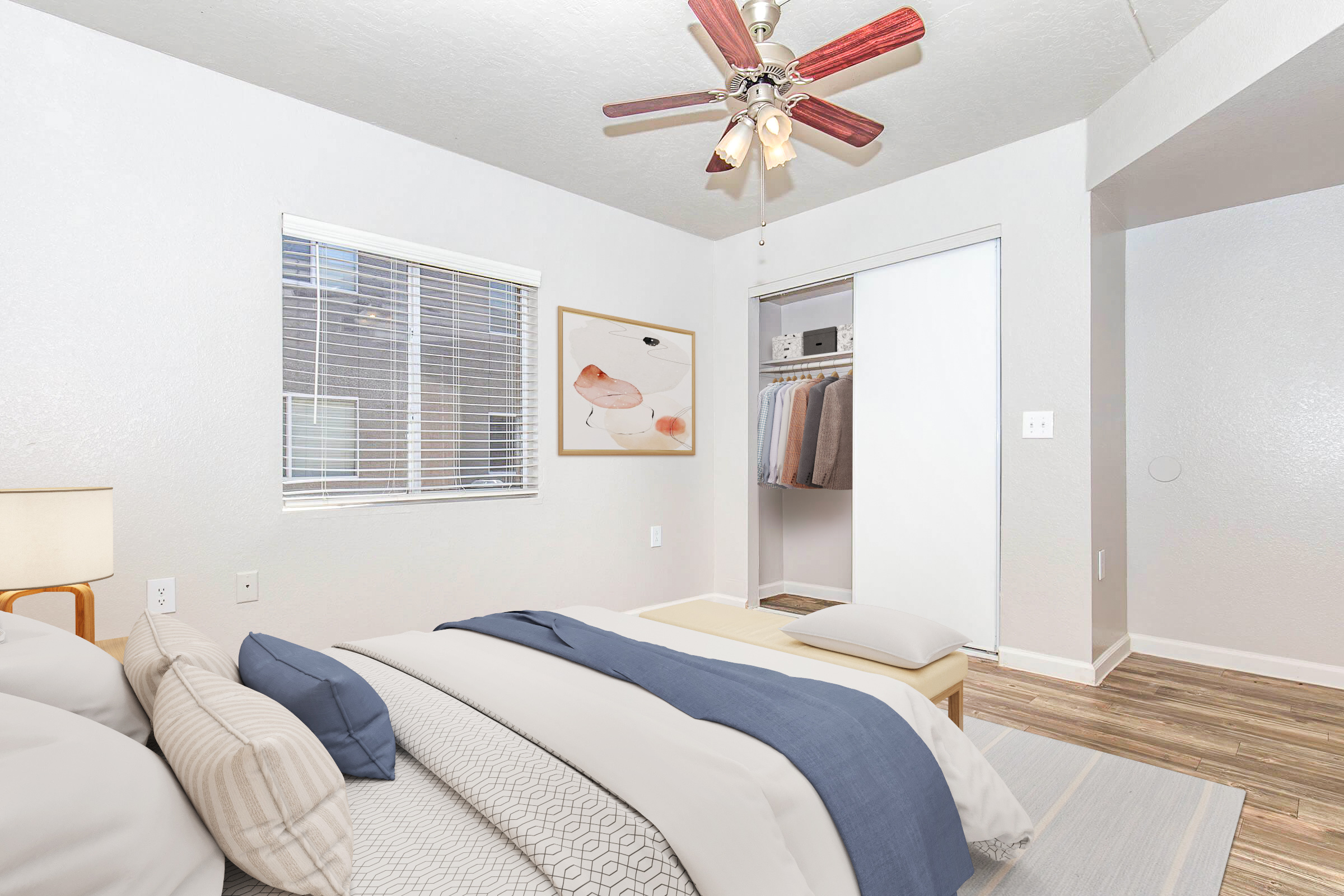 Cozy bedroom with a bed featuring layered bedding in shades of gray and blue. A ceiling fan with wooden blades hangs above, and a framed artwork is mounted on the light-colored wall. Natural light enters through a window, and a closet with neatly arranged clothing is visible.