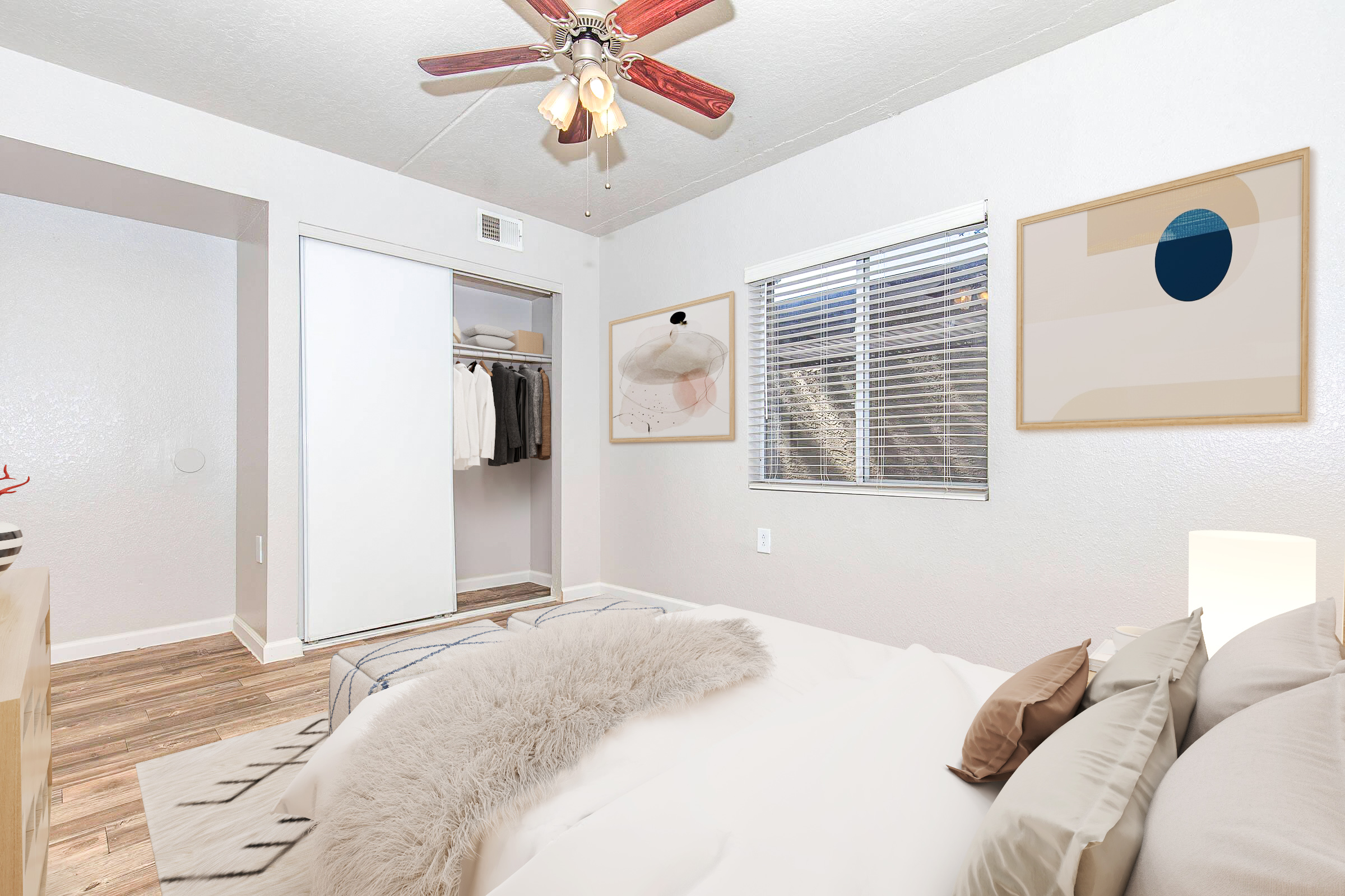 A cozy bedroom featuring a neatly made bed with decorative pillows, a closet with clothes visible, a window with blinds, and modern artwork on the walls. The room has a ceiling fan, wooden flooring, and soft neutral colors, creating a warm and inviting atmosphere.