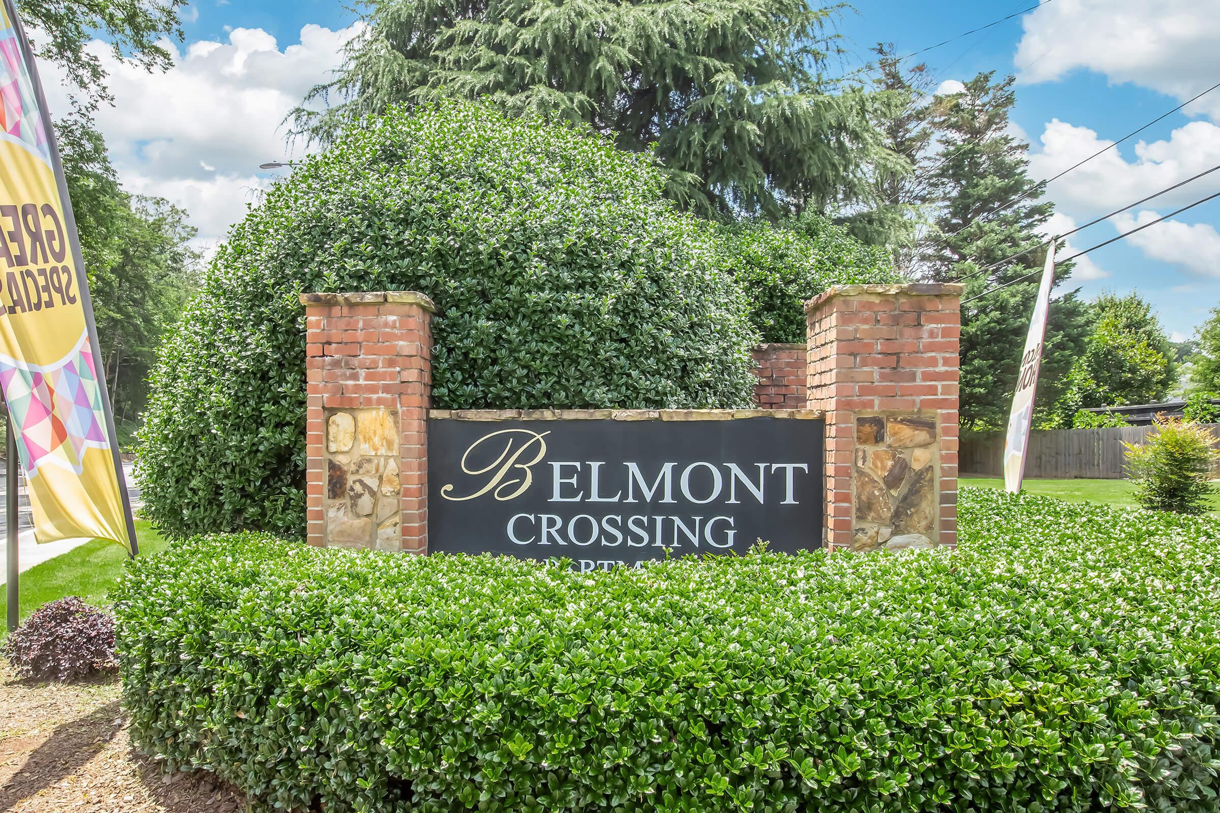 Sign for Belmont Crossing, surrounded by lush greenery and brick pillars. The sign features elegant lettering and is located in a well-maintained area, suggesting a residential or commercial property. Bright sky and trees are visible in the background, enhancing the inviting atmosphere.