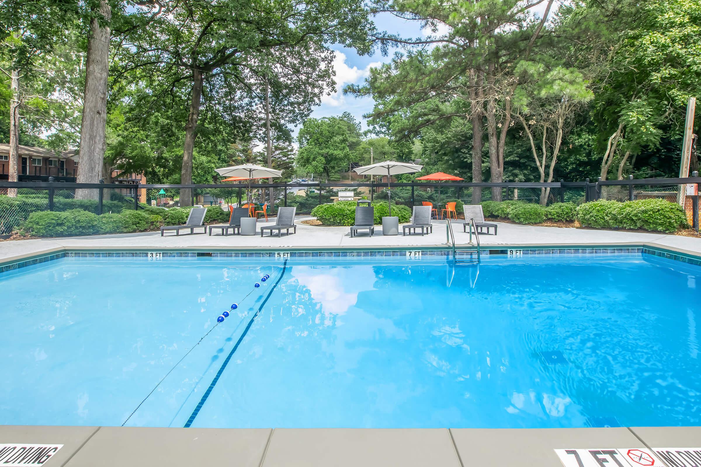 A bright blue pool surrounded by lounge chairs and umbrellas, set in a landscaped outdoor area with trees. The pool area features a fenced perimeter and visible seating, creating a relaxing atmosphere in a residential or resort environment.