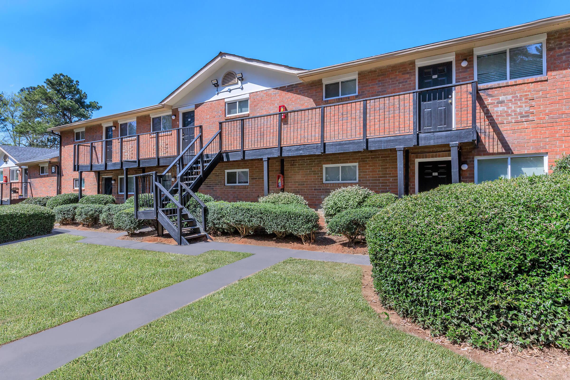 A well-maintained brick apartment complex featuring two levels with black railings, surrounded by neatly trimmed bushes and a green lawn. A paved walkway leads through the grassy area to the entrance of the building, under a clear blue sky.