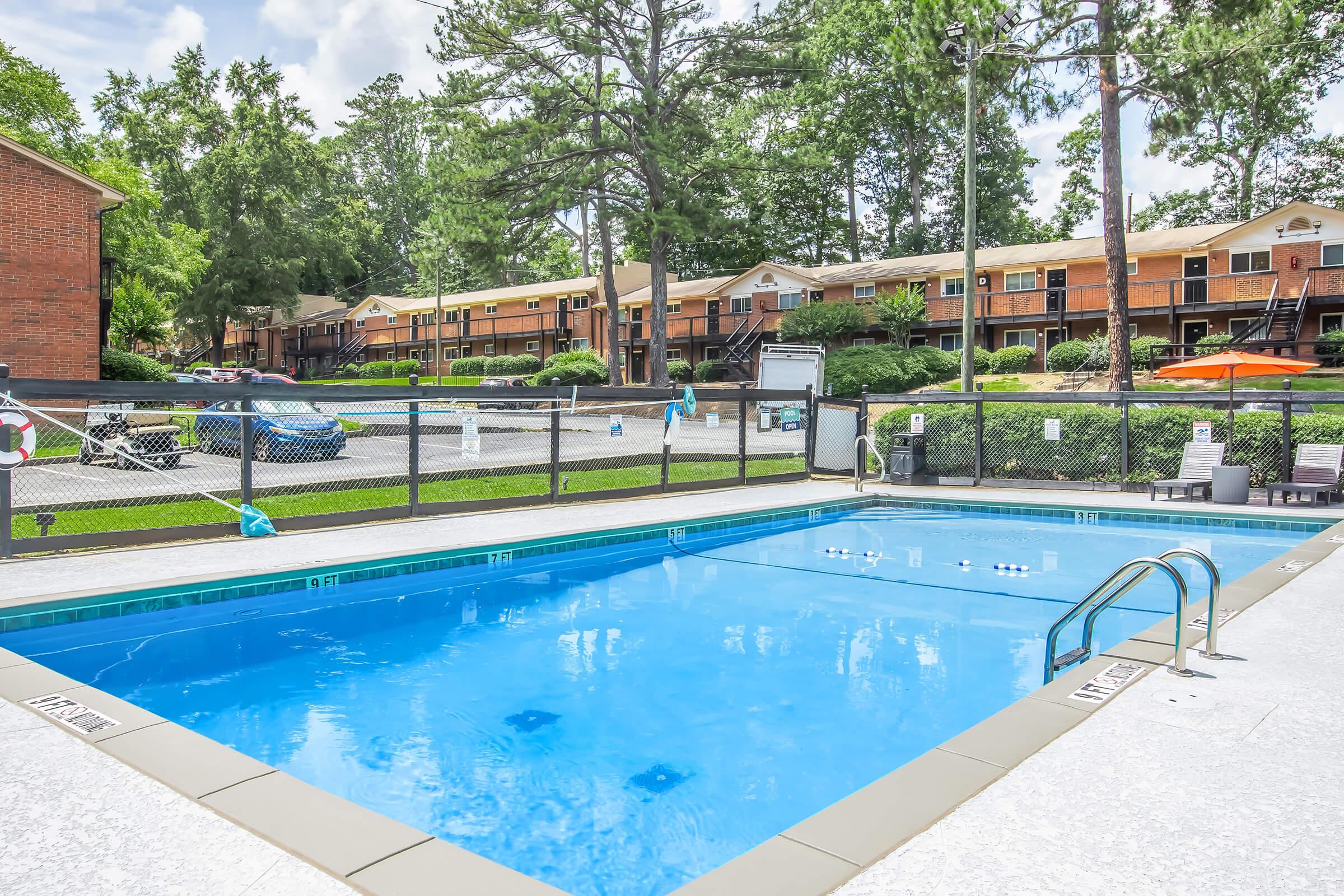 A brightly lit pool area with clear blue water, surrounded by a security fence. In the background, multiple two-story brick apartment buildings are visible, set among trees and green grass. A few lounge chairs and umbrellas are near the pool, suggesting a relaxing outdoor space.