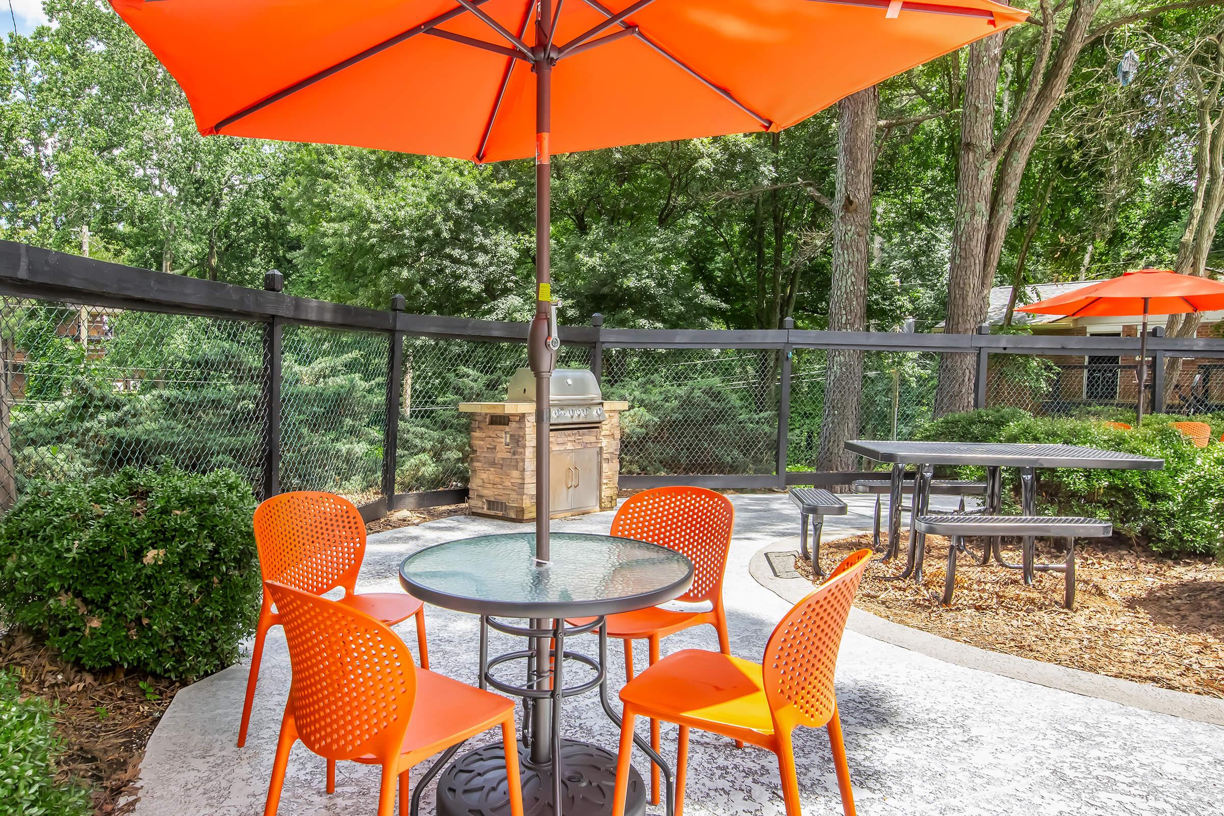 An outdoor seating area featuring a round table surrounded by five orange chairs. An orange umbrella provides shade, and a grill is located in the background near a fence with greenery. The space is designed for leisure and dining in a natural setting.