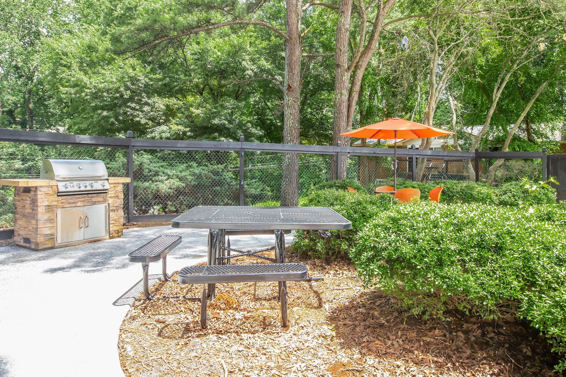 Outdoor patio area featuring a charcoal grill, a black picnic table with benches, and vibrant orange chairs under an umbrella. Surrounded by lush greenery and a fenced area, the picturesque setting invites relaxation and outdoor dining.
