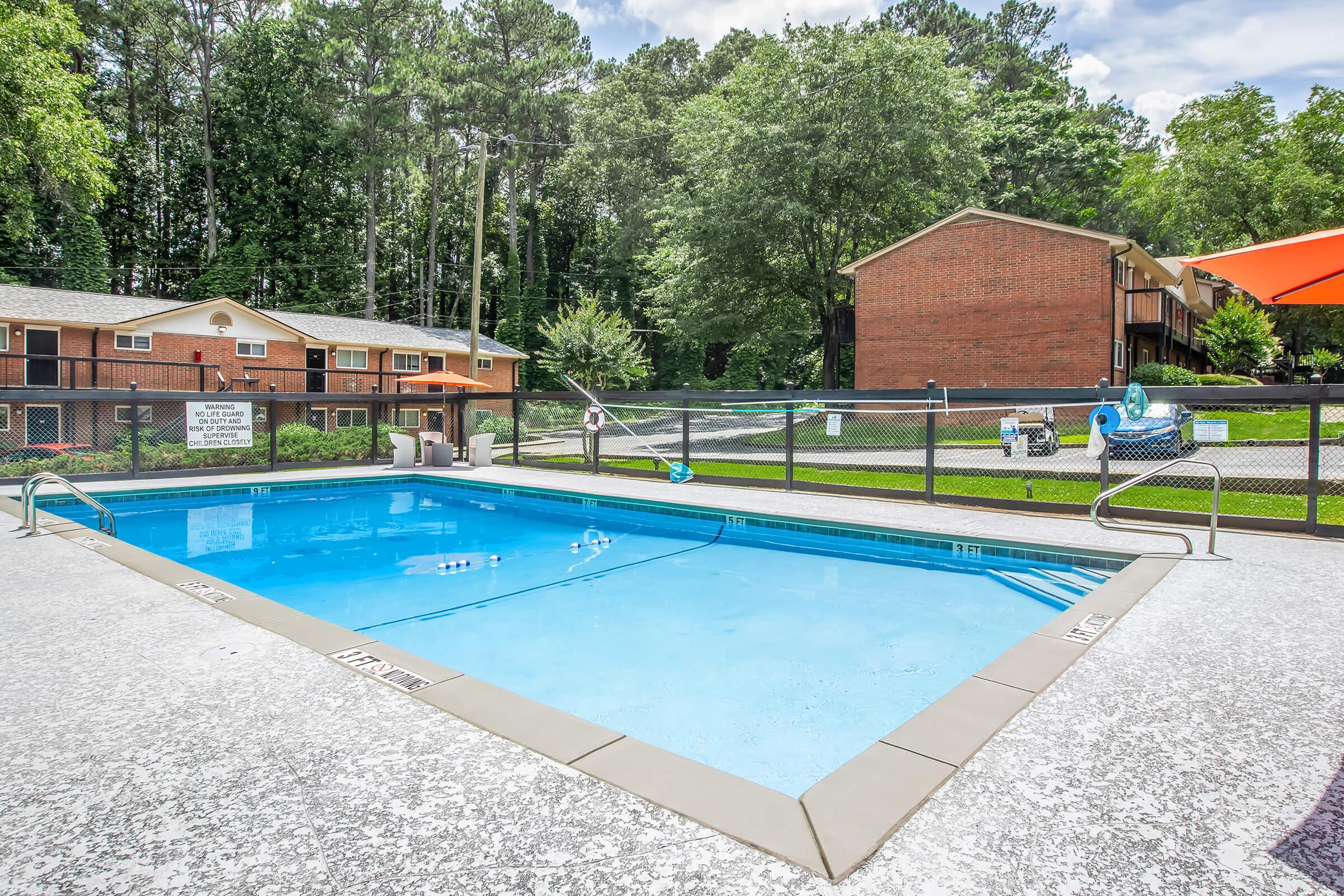 A clear swimming pool surrounded by a fenced area, with lounge chairs nearby. In the background, there are two brick apartment buildings and lush green trees lining the perimeter, under a partly cloudy sky. The area appears well-maintained and inviting for relaxation and recreation.