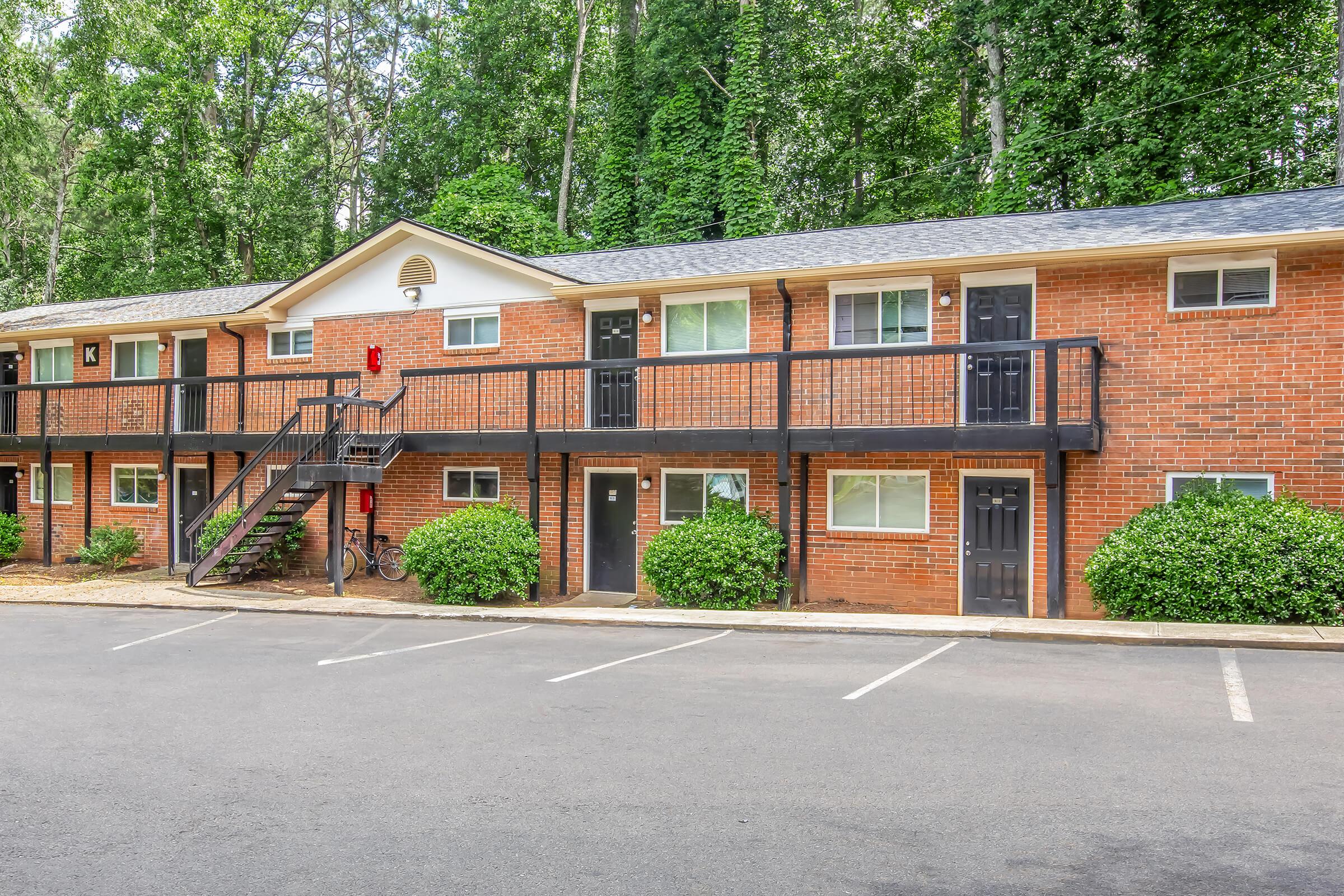 Row of brick apartments with two levels, featuring multiple doors and small balconies. Surrounded by green shrubs and trees in a natural setting. A bicycle is leaned against the railing of the staircase. The parking lot in front has several empty spaces.