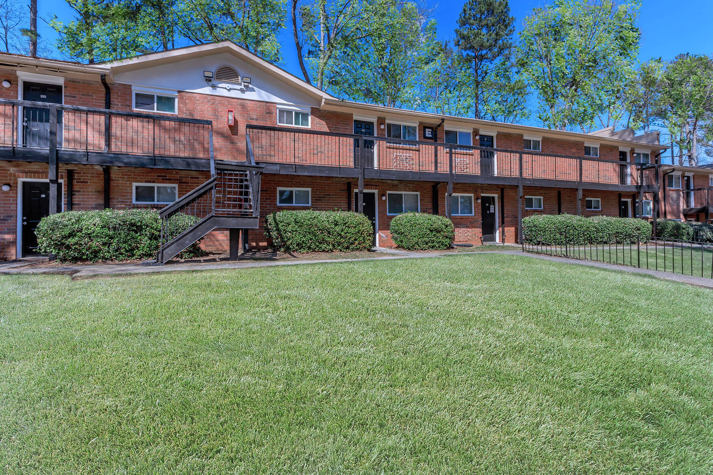 A view of a two-story brick apartment building with balconies, surrounded by well-maintained green grass and shrubs. Trees in the background provide shade under a clear blue sky. The scene conveys a peaceful residential area.
